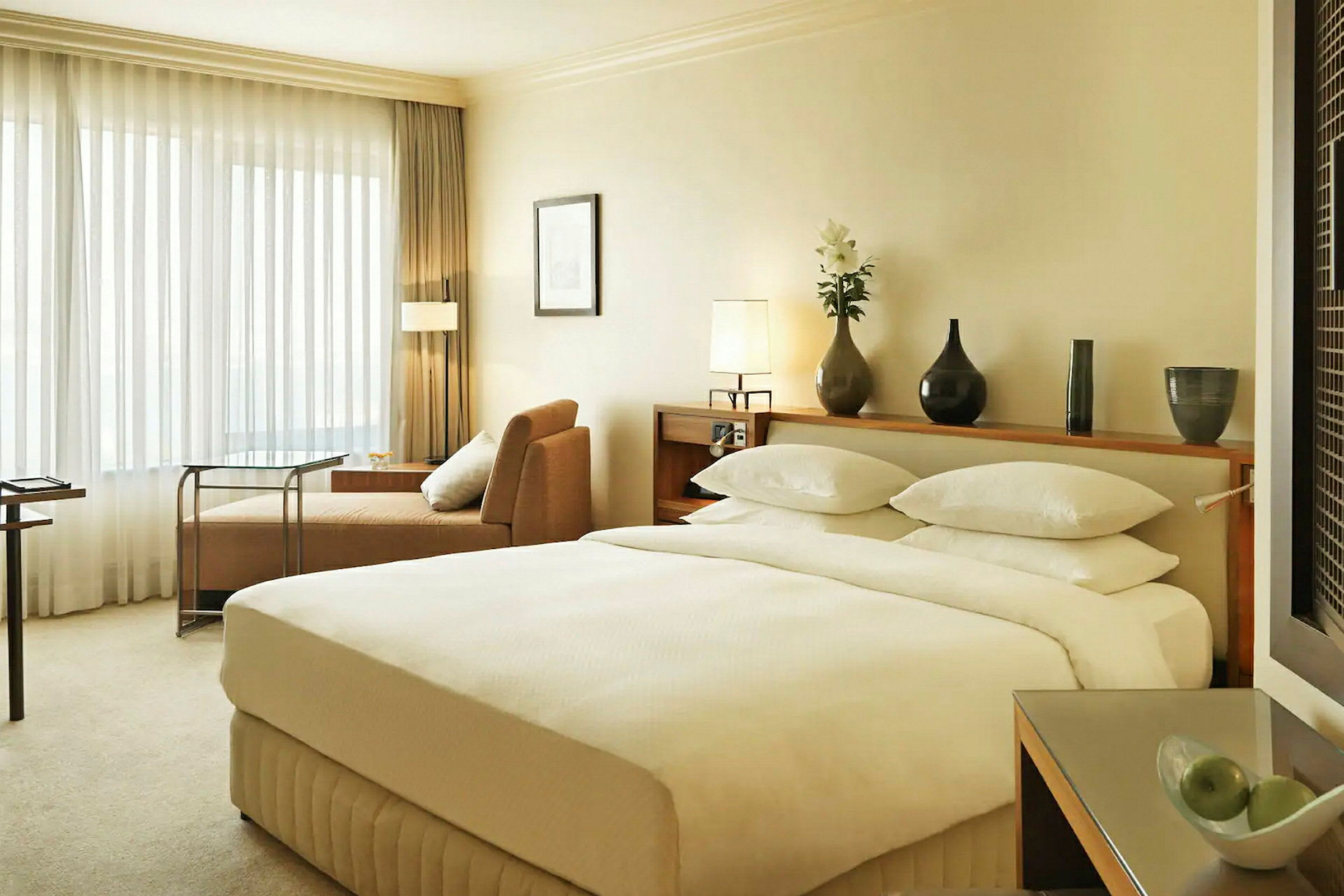 Sovrum i hotellrum med en stor dubbelsäng, beige soffa och ett bord nära ett fönster med genomskinliga gardiner. (AI-genererad text)