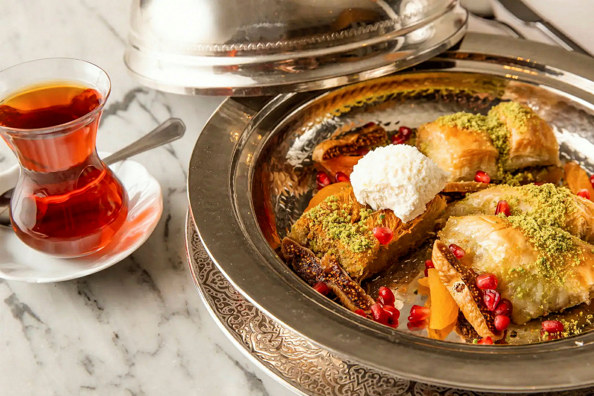 En tallrik med efterrätt, bestående av baklava, pistagenötter, granatäpplefrön och vispad grädde, bredvid ett glas te. (AI-genererad text)