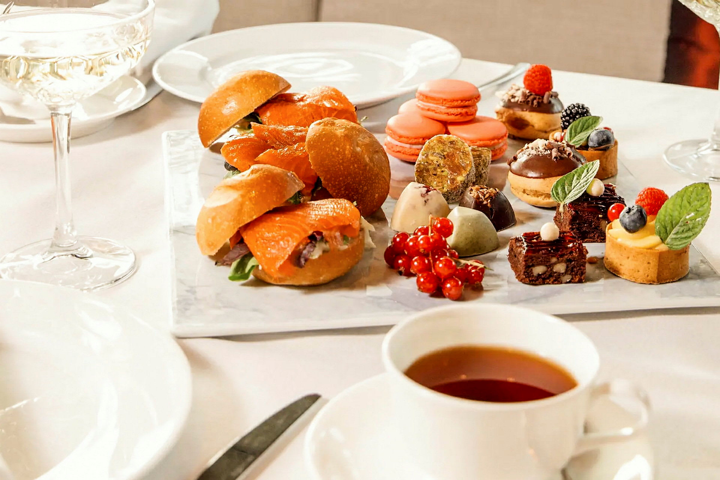 Ett bord med diverse smårätter och desserter, inklusive smörgåsar, macarons och små bakverk. En kopp te och ett glas vin är också framställda. (AI-genererad text)