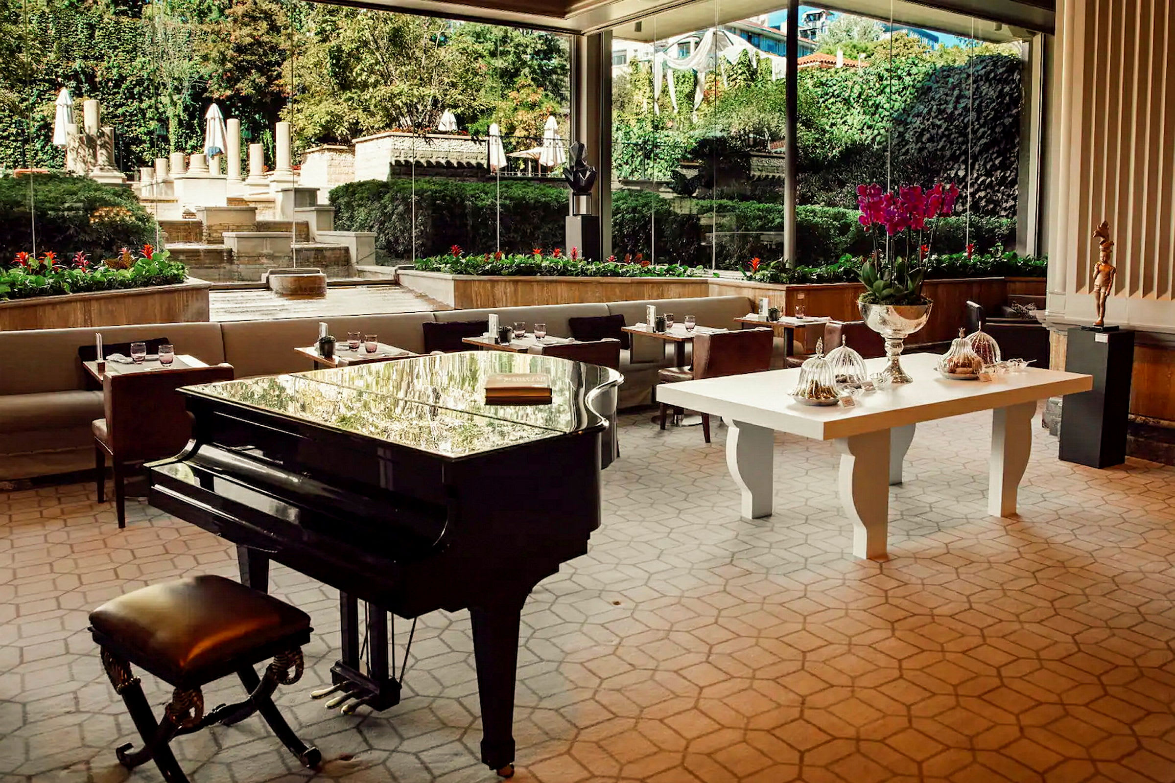 En hotellrestaurang med ett stort svart piano i förgrunden. I bakgrunden finns soffor, bord och utsikt över en trädgård med vegetation och trappor. (AI-genererad text)