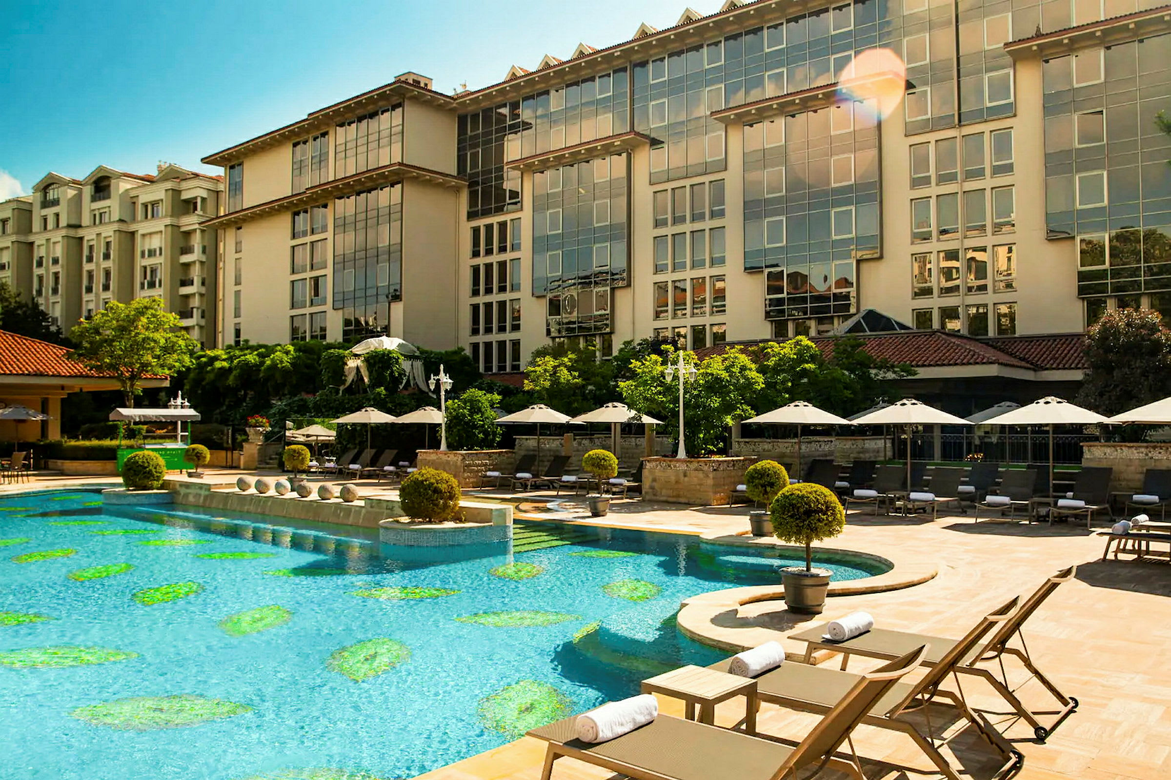 En utomhuspool vid ett hotell, omgiven av solstolar och parasoll. Hotellets byggnad syns i bakgrunden. (AI-genererad text)