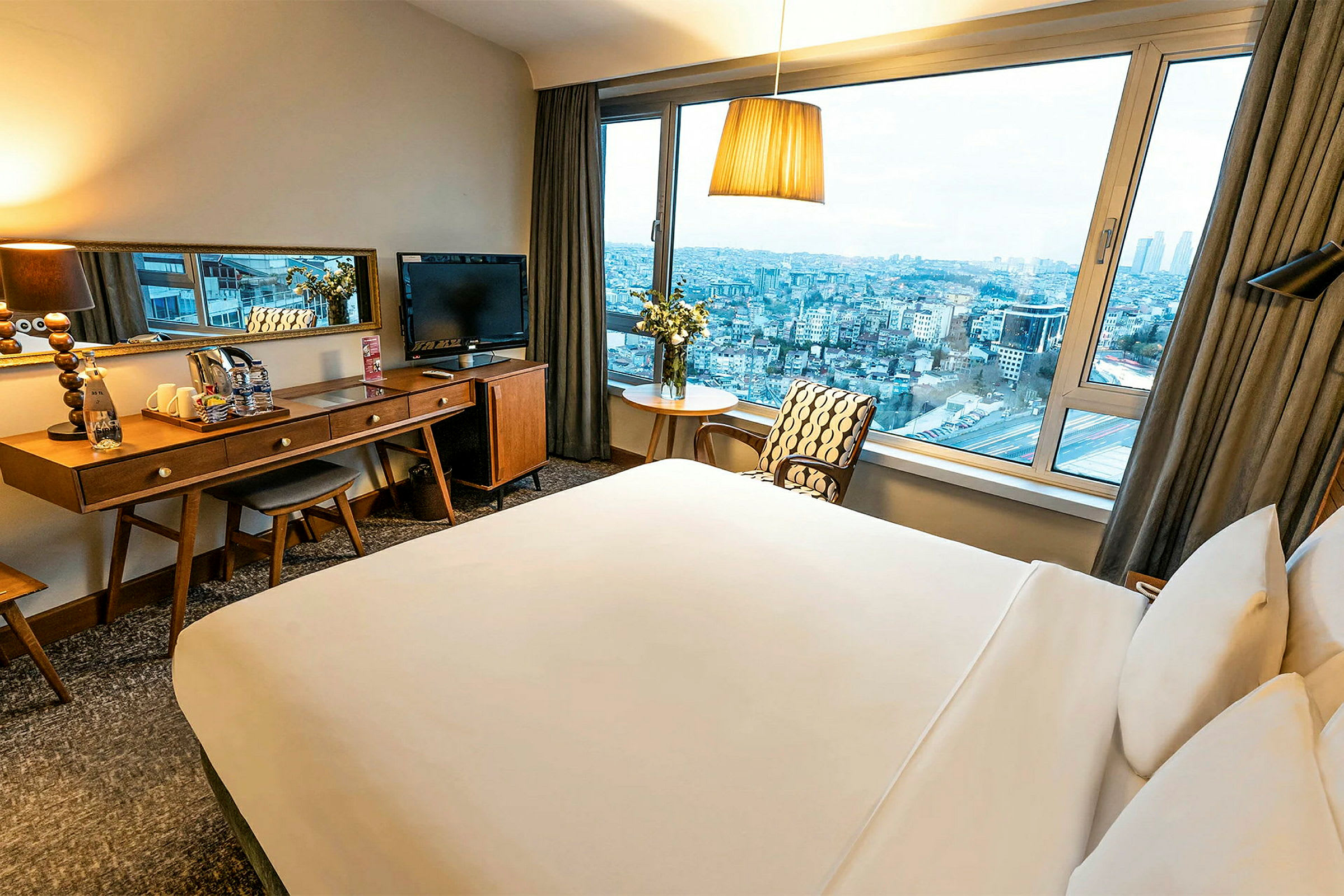 Ett hotellrum med en stor säng, skrivbord och spegel samt en stor fönsterutsikt över en stad. (AI-genererad text)