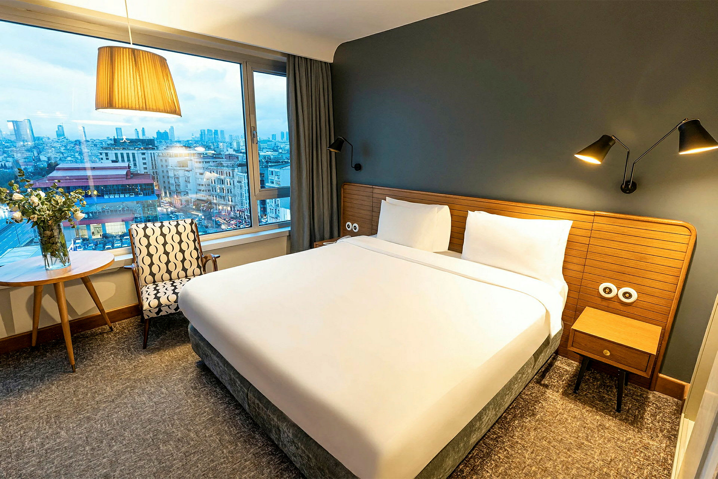 Ett hotellrum med en stor säng, fönster och utsikt över en stad. (AI-genererad text)