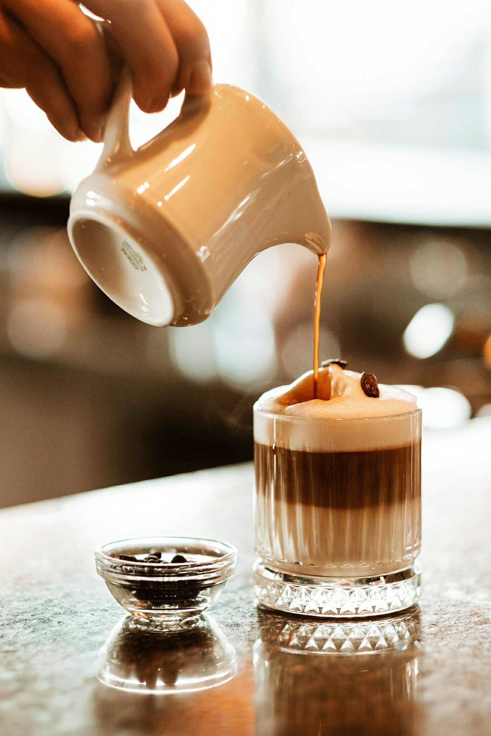 En hand häller en varm dryck, möjligtvis kaffe, från en kanna ner i ett glas med skum på toppen. Bredvid glaset står en liten skål med kaffebönor. (AI-genererad text)