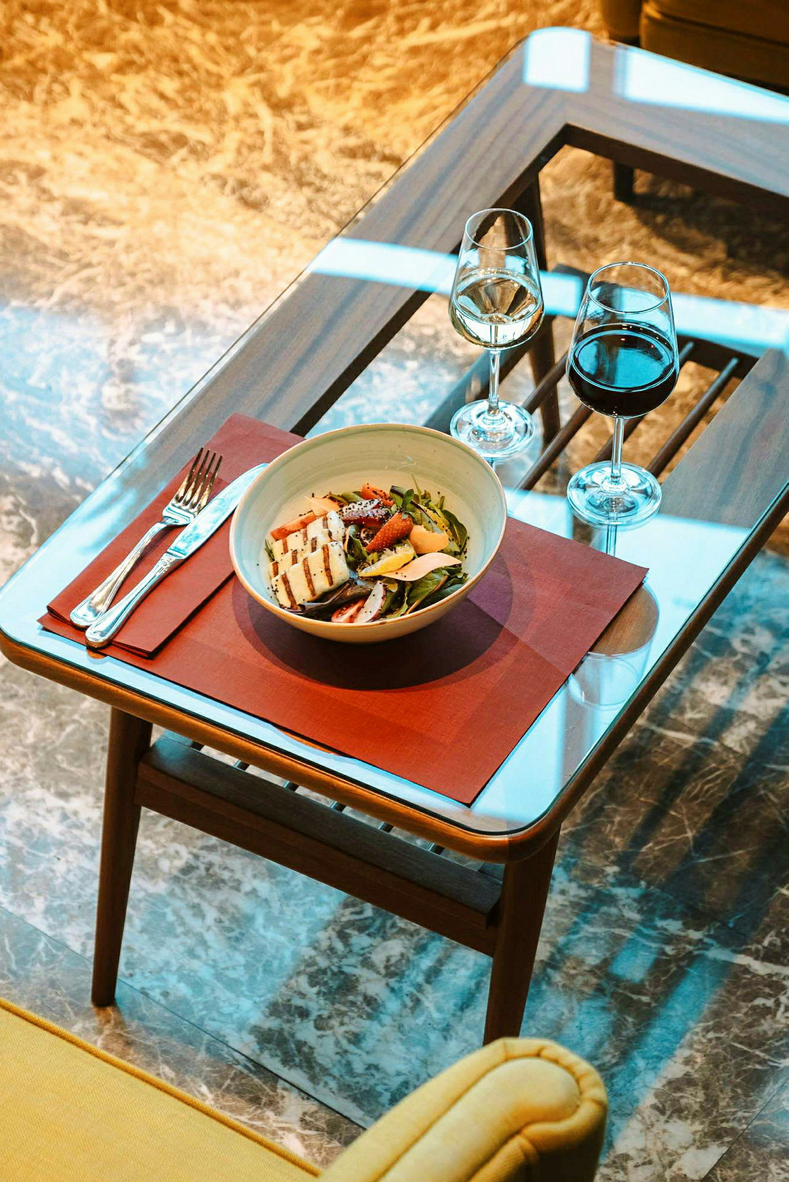 Bilden visar ett glasbord med en salladsskål, bestick och två glas vin placerade på en röd bordstablett. (AI-genererad text)
