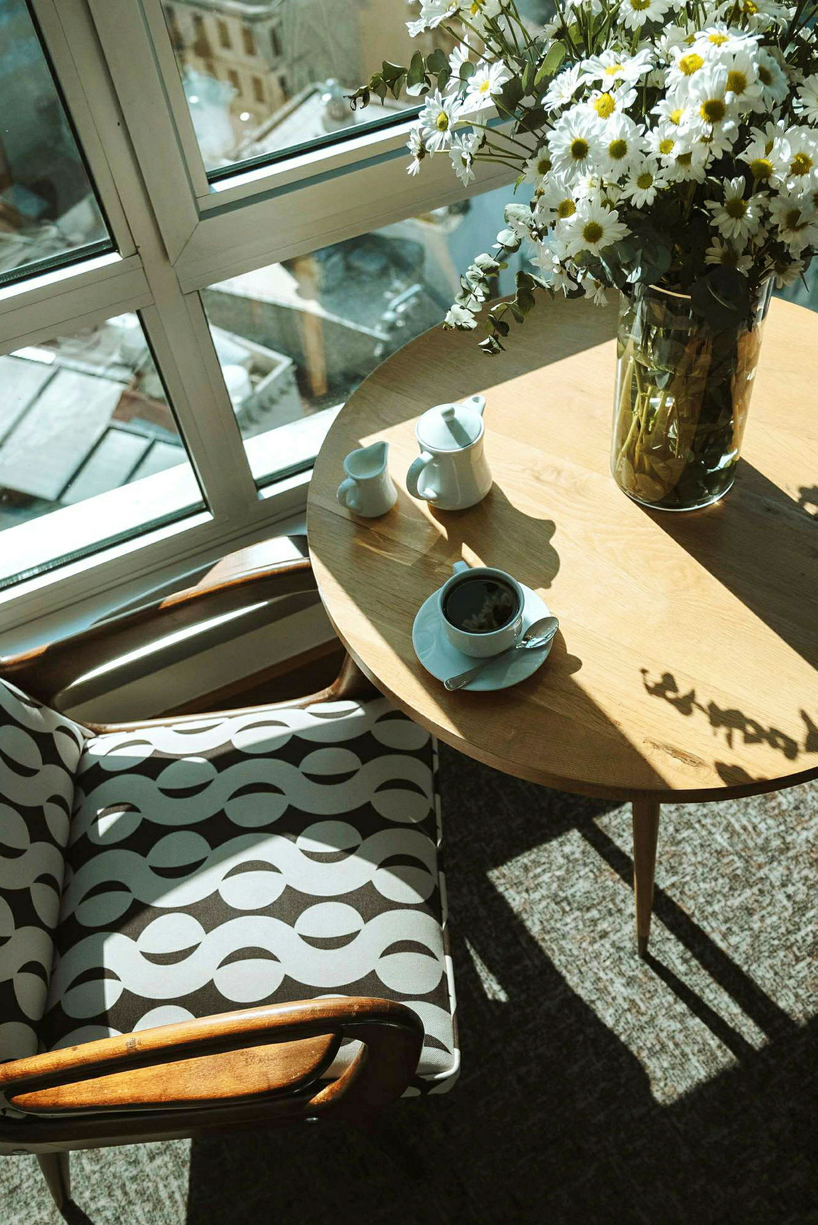 Ett bord med en kaffekopp, oblat och en mjölkkanna intill en stol med mönstrat tyg, sett framför ett stort fönster med utsikt. (AI-genererad text)