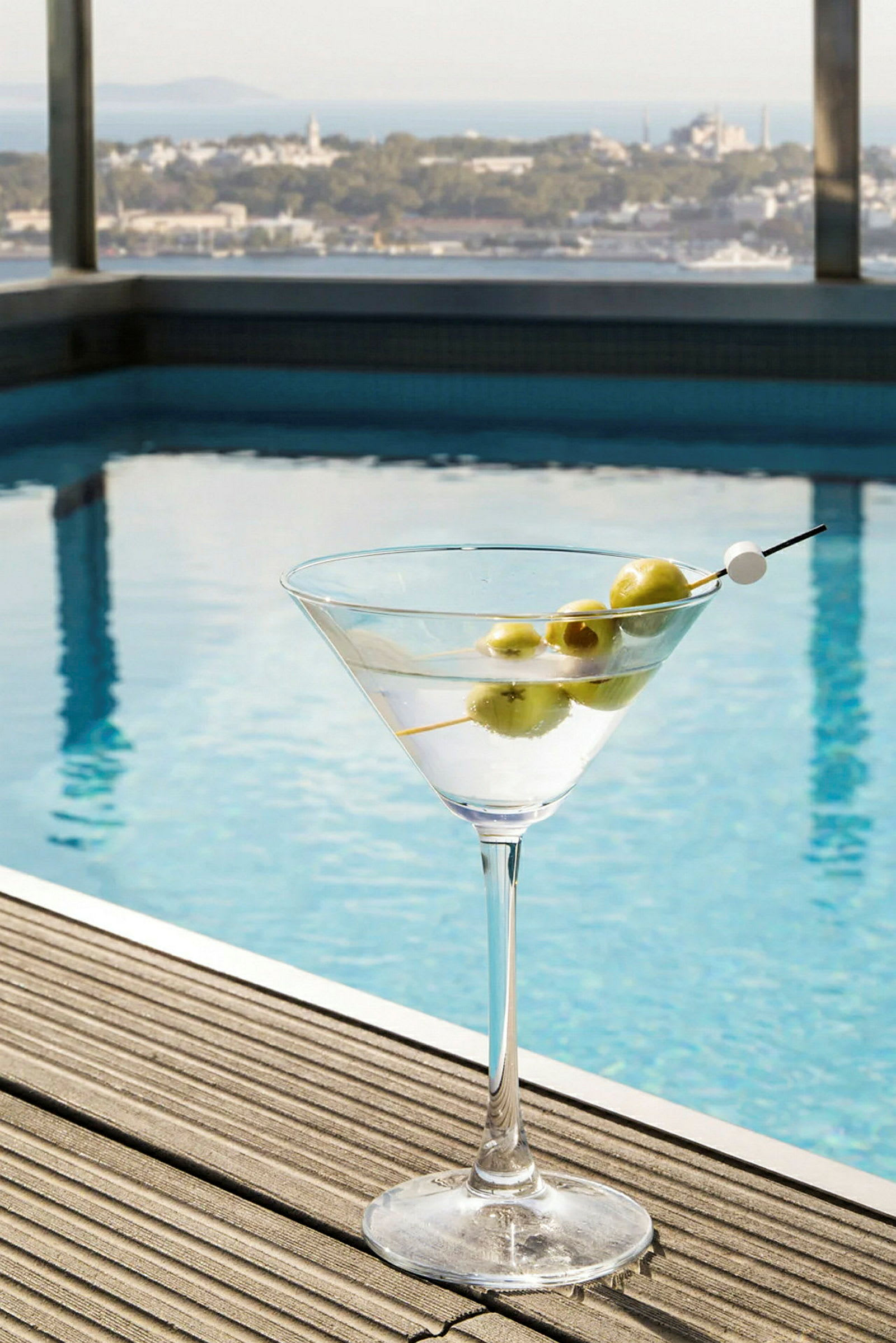 Ett glas med en cocktail och oliver på en träyta bredvid en pool, med en utsikt över en stadslandskap. (AI-genererad text)