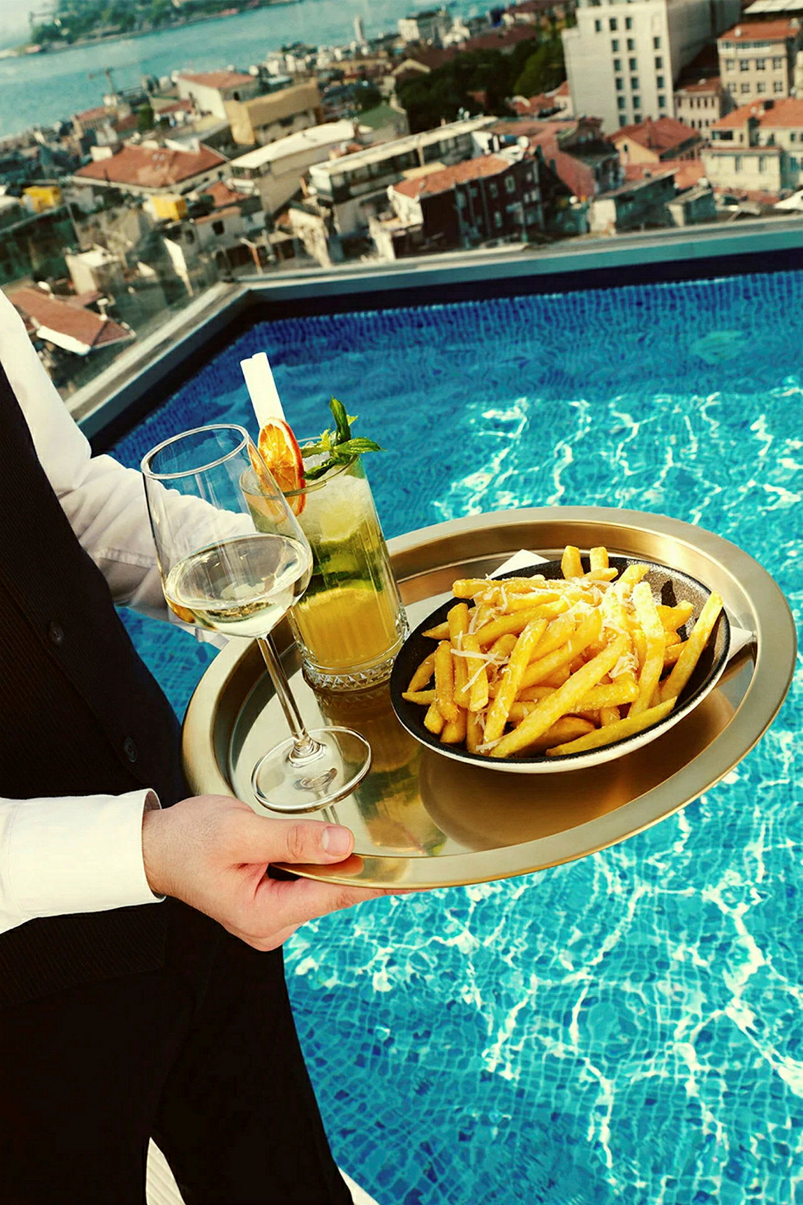 En serveringsbricka med pommes frites på en tallrik, ett glas vitt vin och en cocktail placeras vid en pool med utsikt över en stad. (AI-genererad text)