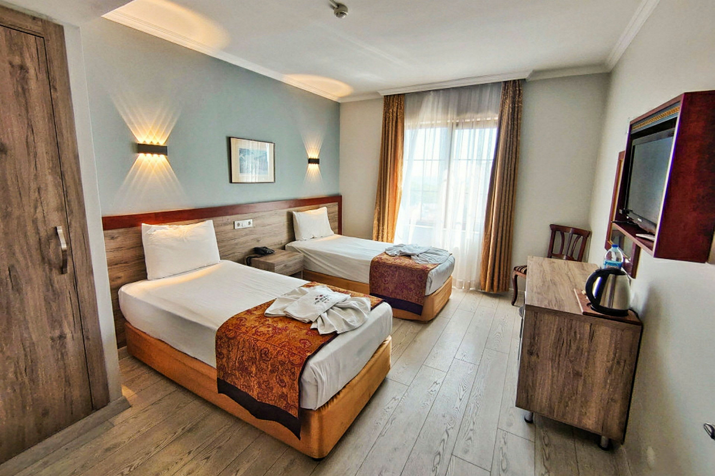 Bild av ett hotellrum med två enkelsängar, ett skrivbord med vattenkokare och en vägg-TV. Rummet har ljusa gardiner och trägolv. (AI-genererad text)