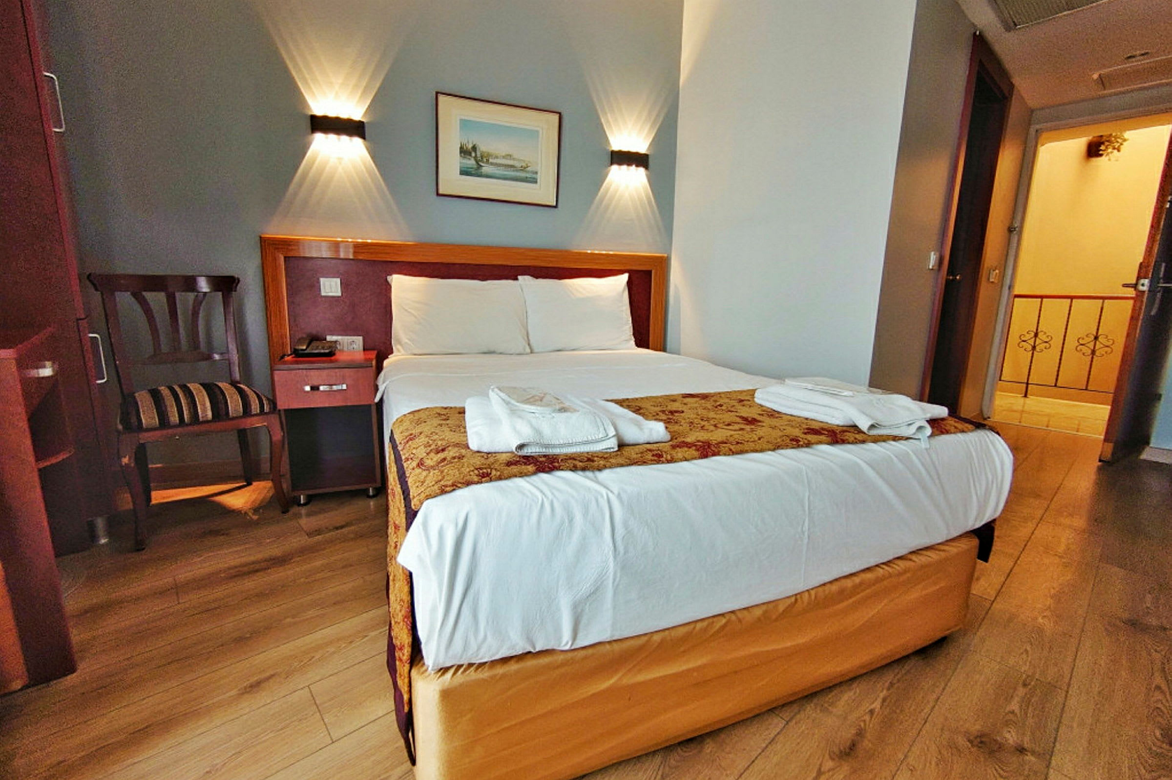 Ett hotellrum med en dubbelsäng, sänglampor på väggen, en stol och bord bredvid sängen, samt öppning mot en hall. (AI-genererad text)