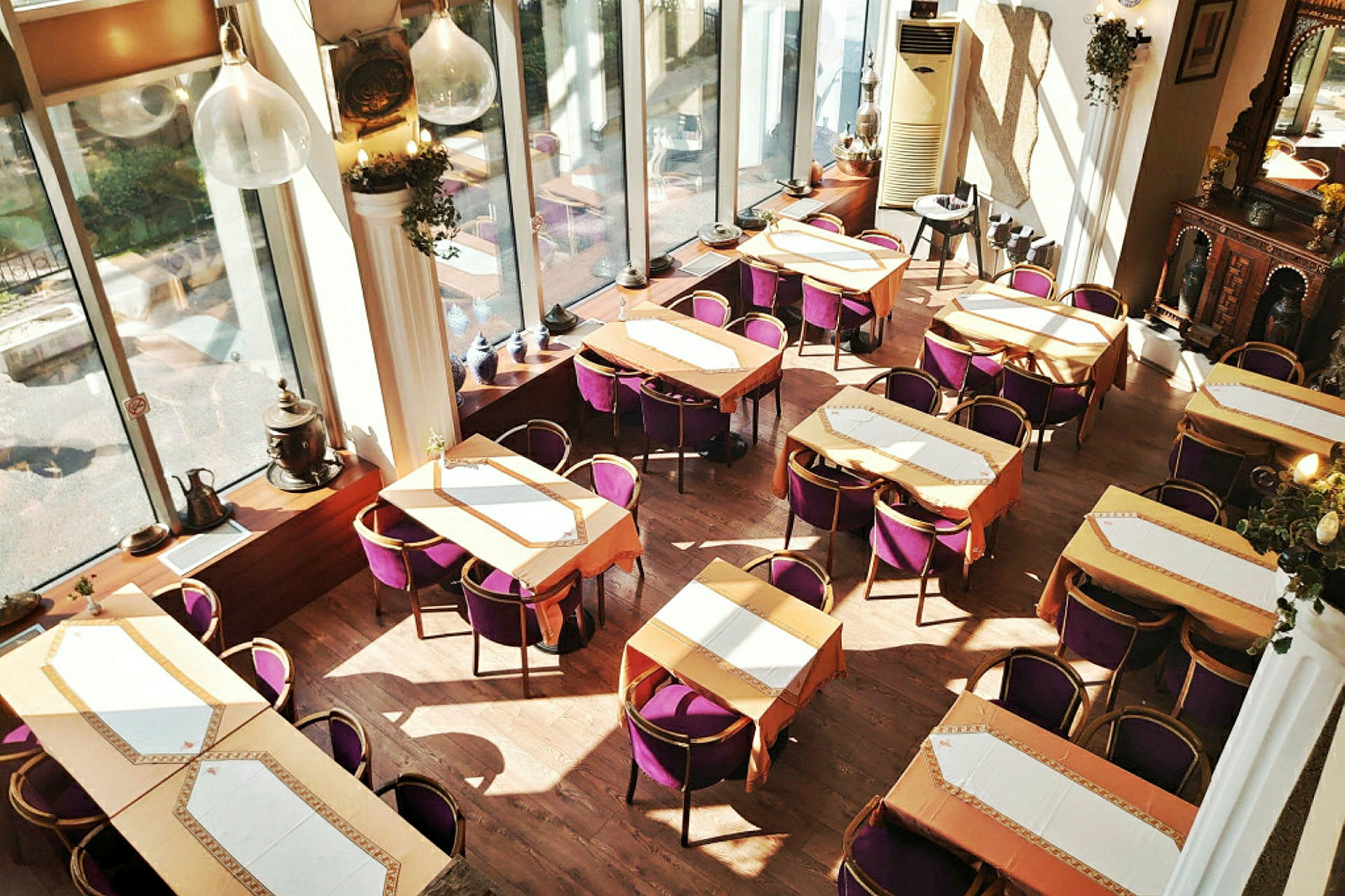 Interiör av en restaurang med flera bord och lila stolar, stora fönster och dekorativa element. (AI-genererad text)