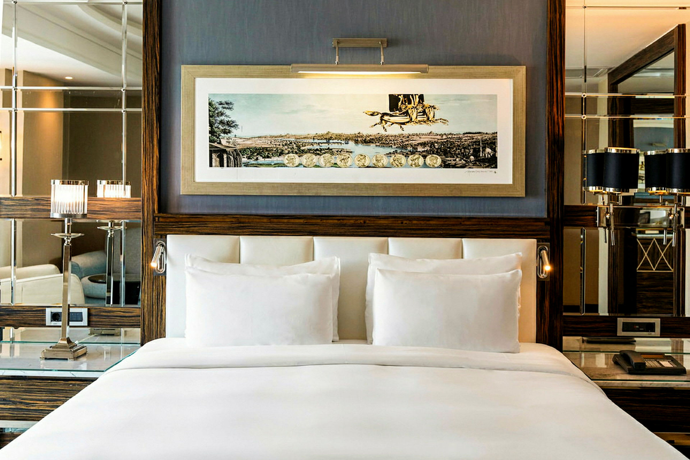 En elegant hotellrumsinteriör med en bred, vitbäddad säng framför en vägg dekorerad med en inramad konstbild, speglar och eleganta lampor. (AI-genererad text)