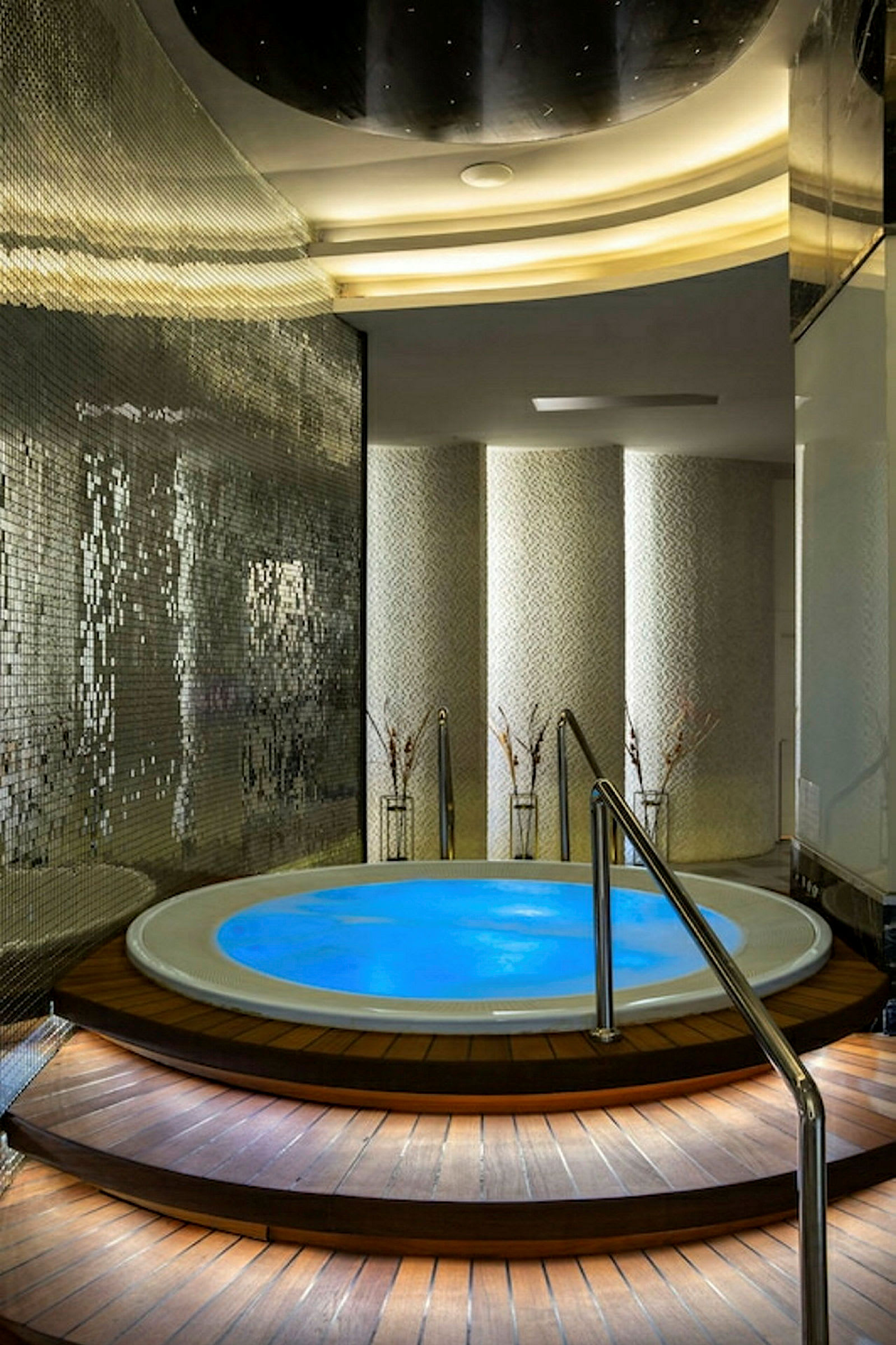 Ett spaområde med en rund jacuzzi omgiven av träpaneler och metallräcken. Väggarna har detaljer av mosaik och belysta inredningselement. (AI-genererad text)