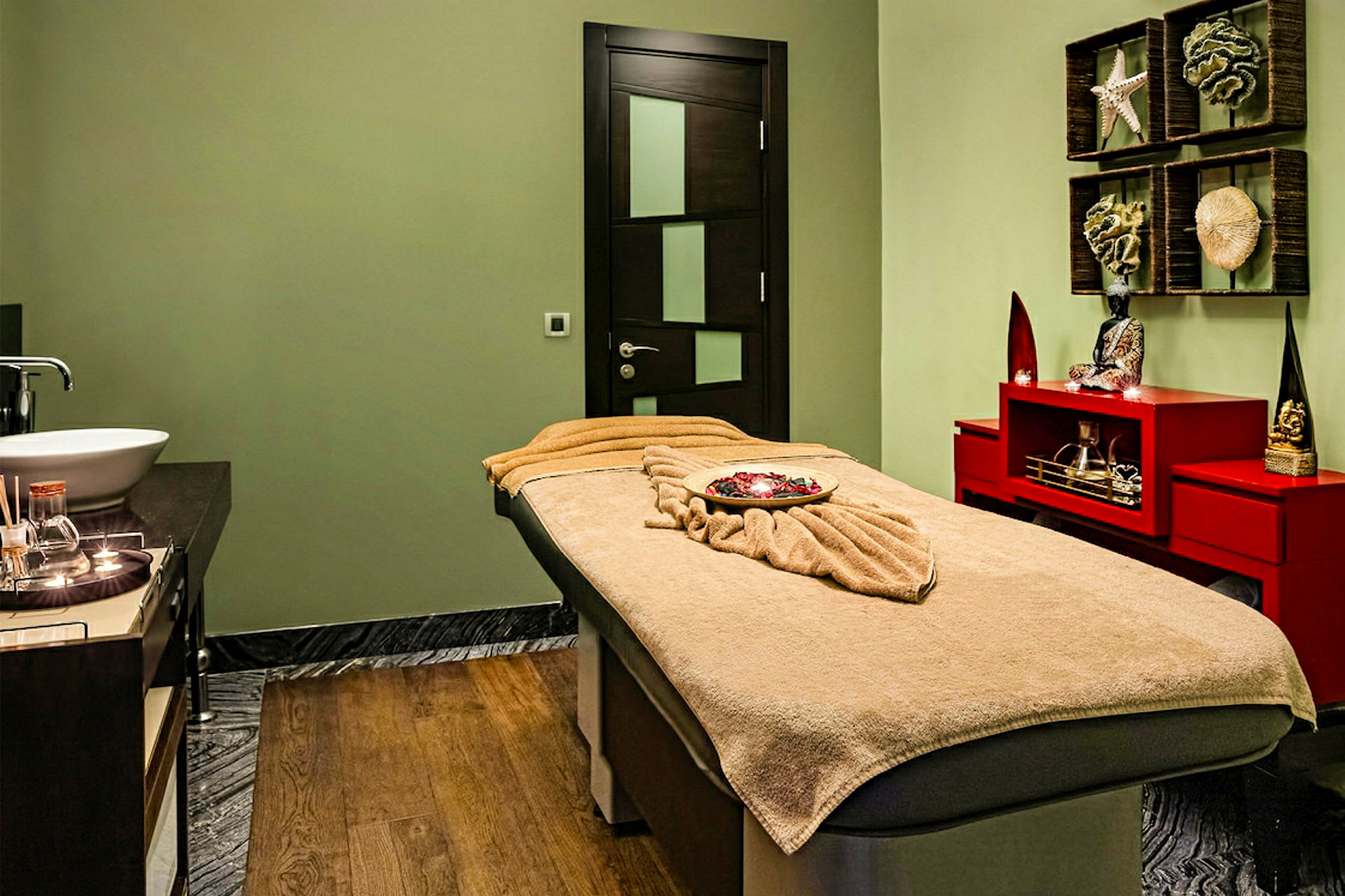 Ett behandlingsrum med en massagebänk täckt av en beige handduk, en vask och en hylla med dekorativa utsmyckningar. (AI-genererad text)
