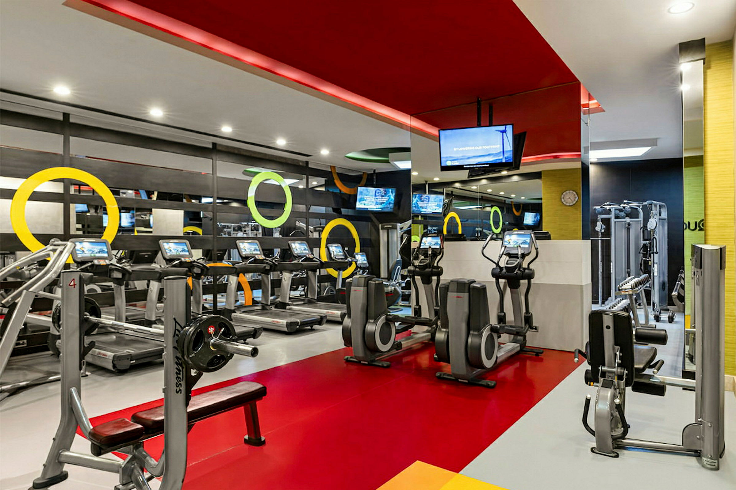 En bild av ett gym med olika träningsutrustningar, såsom löpband, crosstrainers och andra maskiner, samt färgglad inredning. (AI-genererad text)