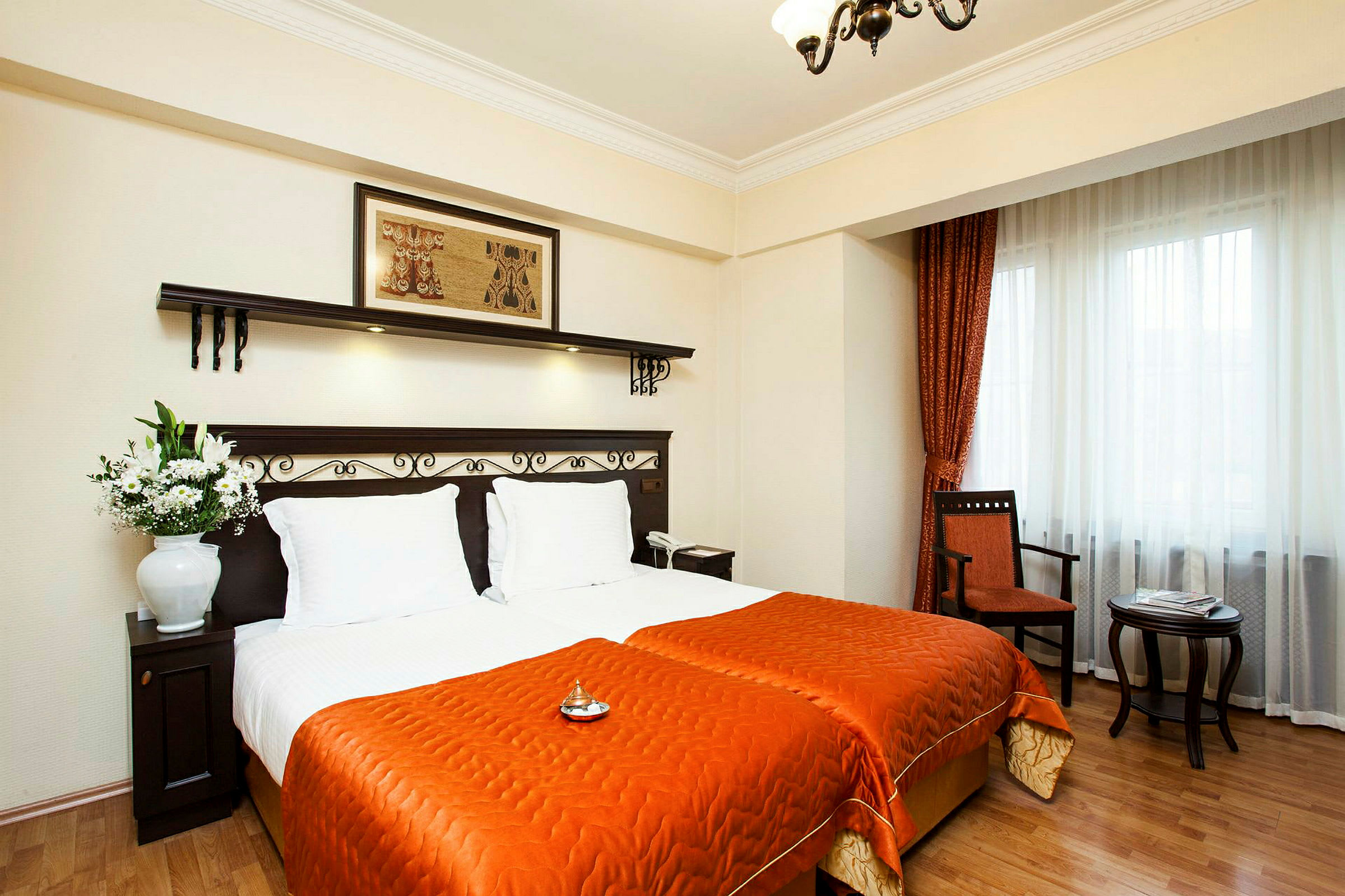 Ett hotellrum med två enkelsängar täckta med orange sängkläder. Sängarna har vita kuddar och ett mörkt trähuvudgavel med dekorativa detaljer. En blomvas står på ett mörkt träbord bredvid sängarna. Fönstret har vita gardiner och orange draperier, och nära det finns en stol och ett litet sidobord. (AI-genererad text)
