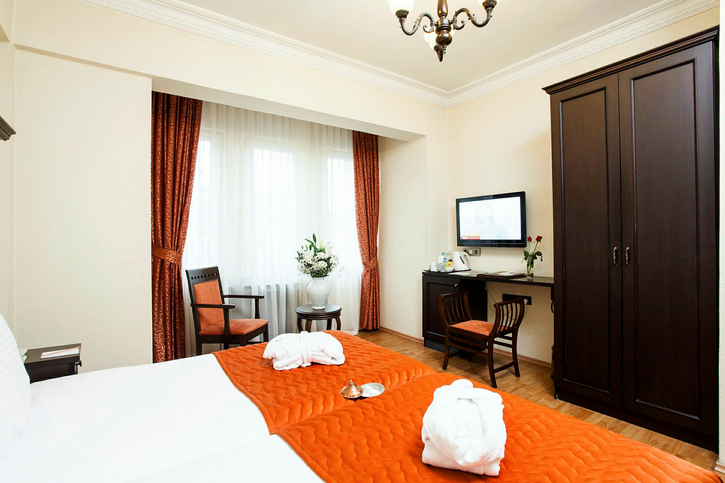 Ett hotellrum med en dubbelsäng bäddad med orange sängkläder. Rummet har mörka möbler, inklusive en garderob, ett skrivbord med en stol, och en väggmonterad TV. Fönstret har vita gardiner med oranga draperier. (AI-genererad text)