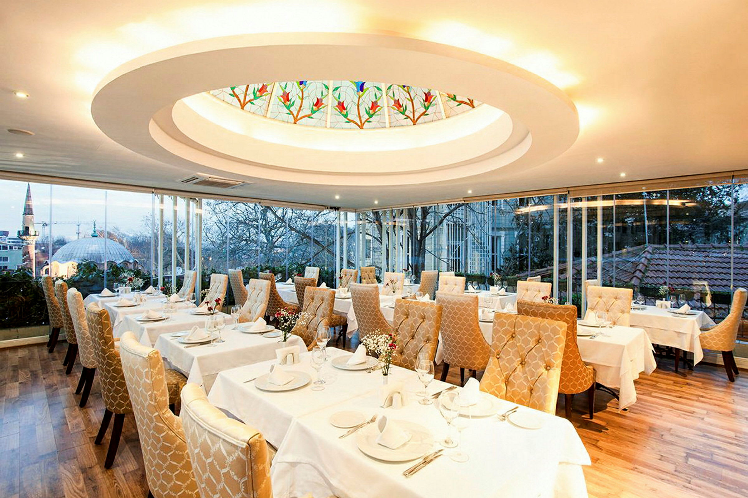 En elegant restaurang med dukade bord och gyllene vadderade stolar. Ett stort fönster visar utsikten över en stad med träd och byggnader. (AI-genererad text)