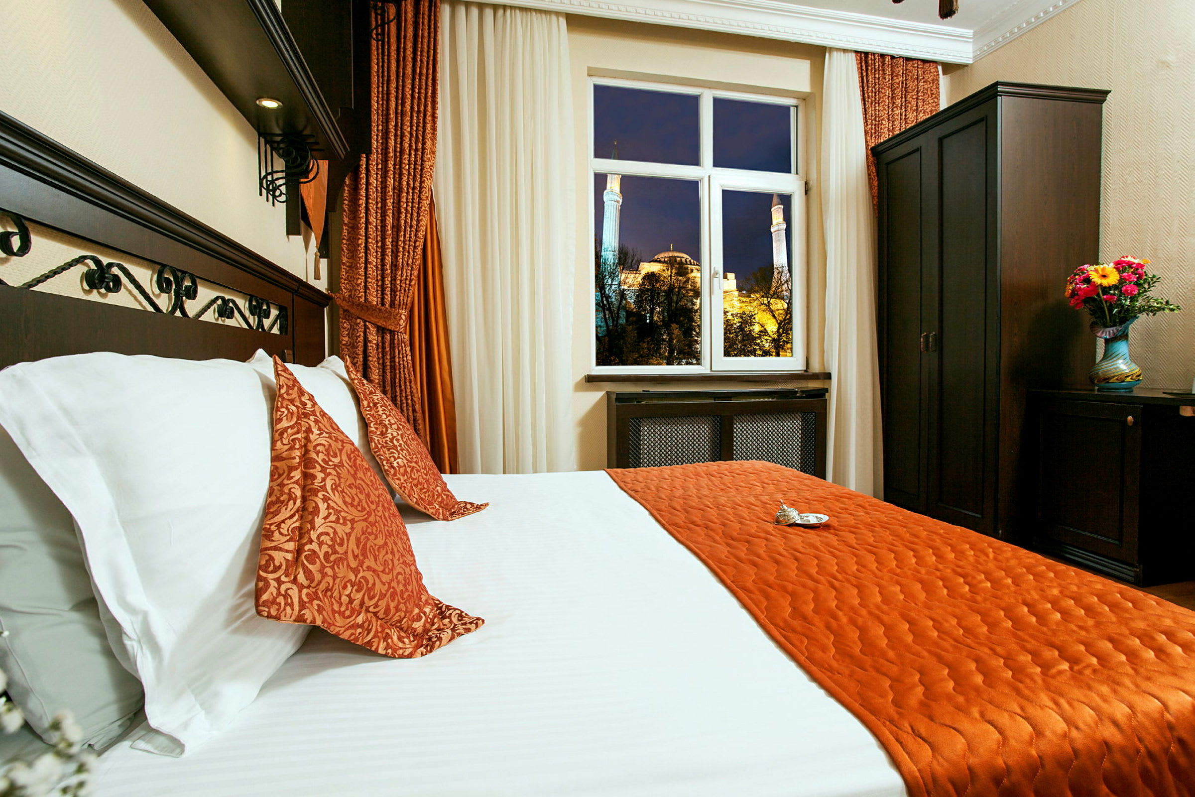 Ett hotellrum med en säng klädd i vita och orangefärgade sängkläder. Ett fönster visar en kvällsvy av Hagia Sofia utanför. Mörka möbler och en vas med blommor syns i rummet. (AI-genererad text)