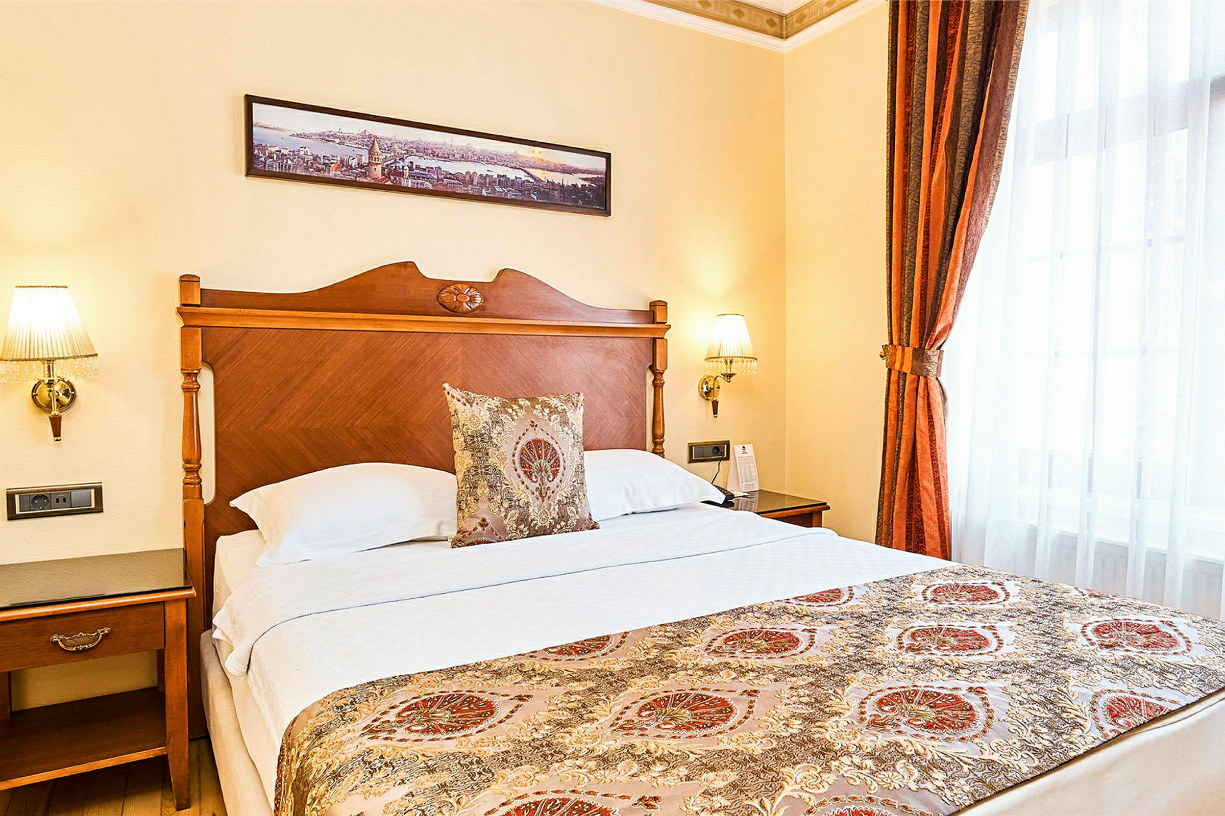Hotellrum med en dubbelsäng och eleganta dekorationer. Sänggaveln är i trä, och sängen har en dekorerad överkast samt prydnadskuddar. På väggen ovanför hänger en panorama som konst. (AI-genererad text)