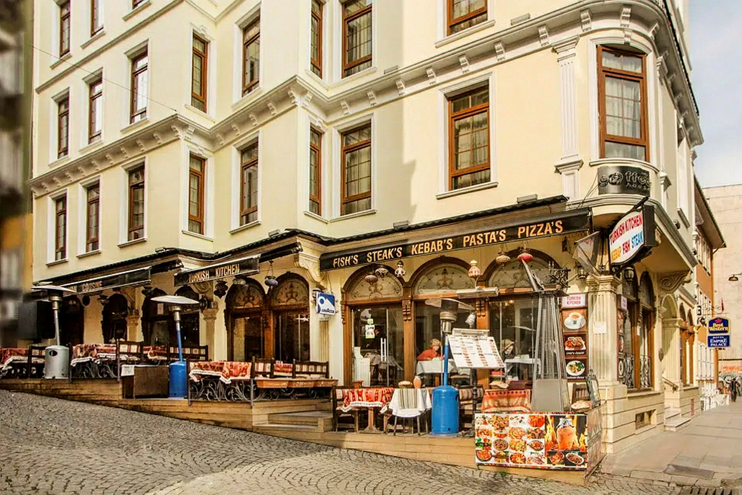 En gata med en byggnad som har restaurang med uteservering, skyltar för mat som kebab och pizza. (AI-genererad text)
