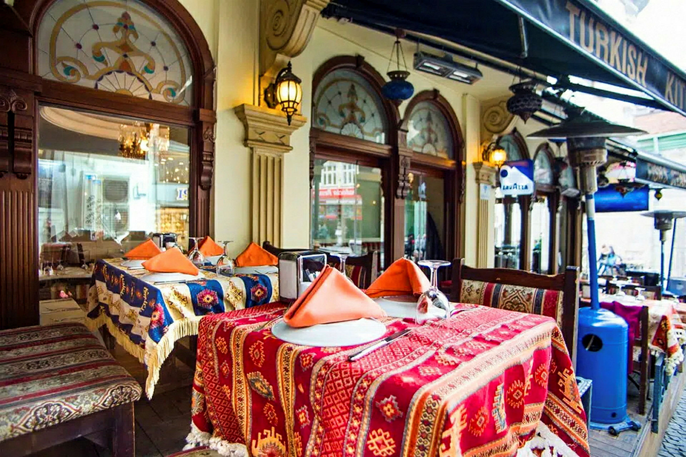 Bord täckta med färgglada tygdukar och orange servetter utanför en restaurang med dekorerade fönster och tak under en baldakin. (AI-genererad text)