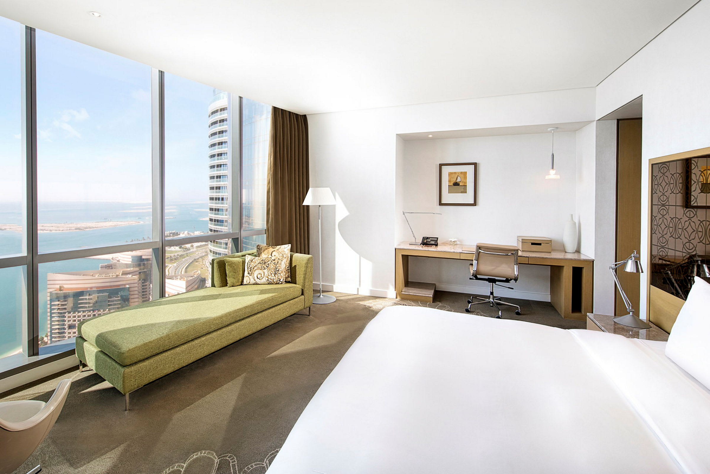 Hotellrum med säng, skrivbord och panoramafönster med utsikt över en kuststad. (AI-genererad text)