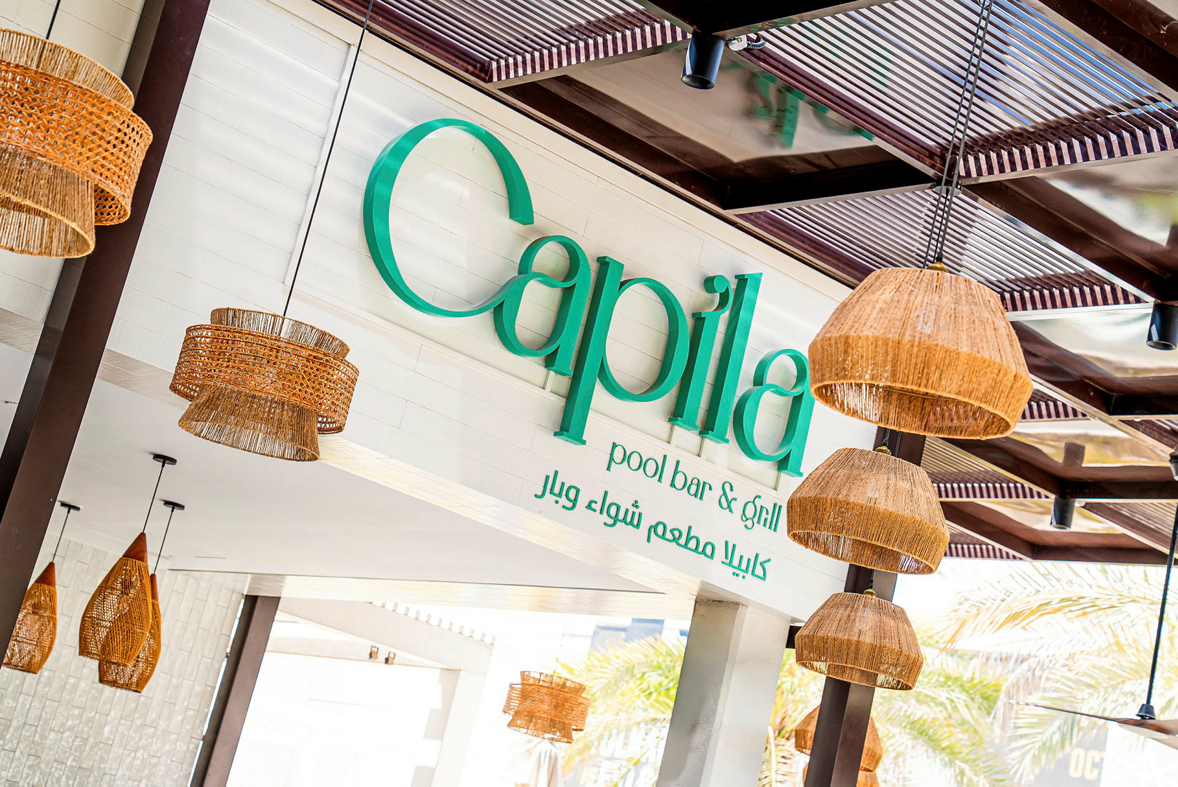 En utomhusskylt med texten 'Capila pool bar & grill' och dekorativa ljuslyktor av naturmaterial hängande från taket. (AI-genererad text)