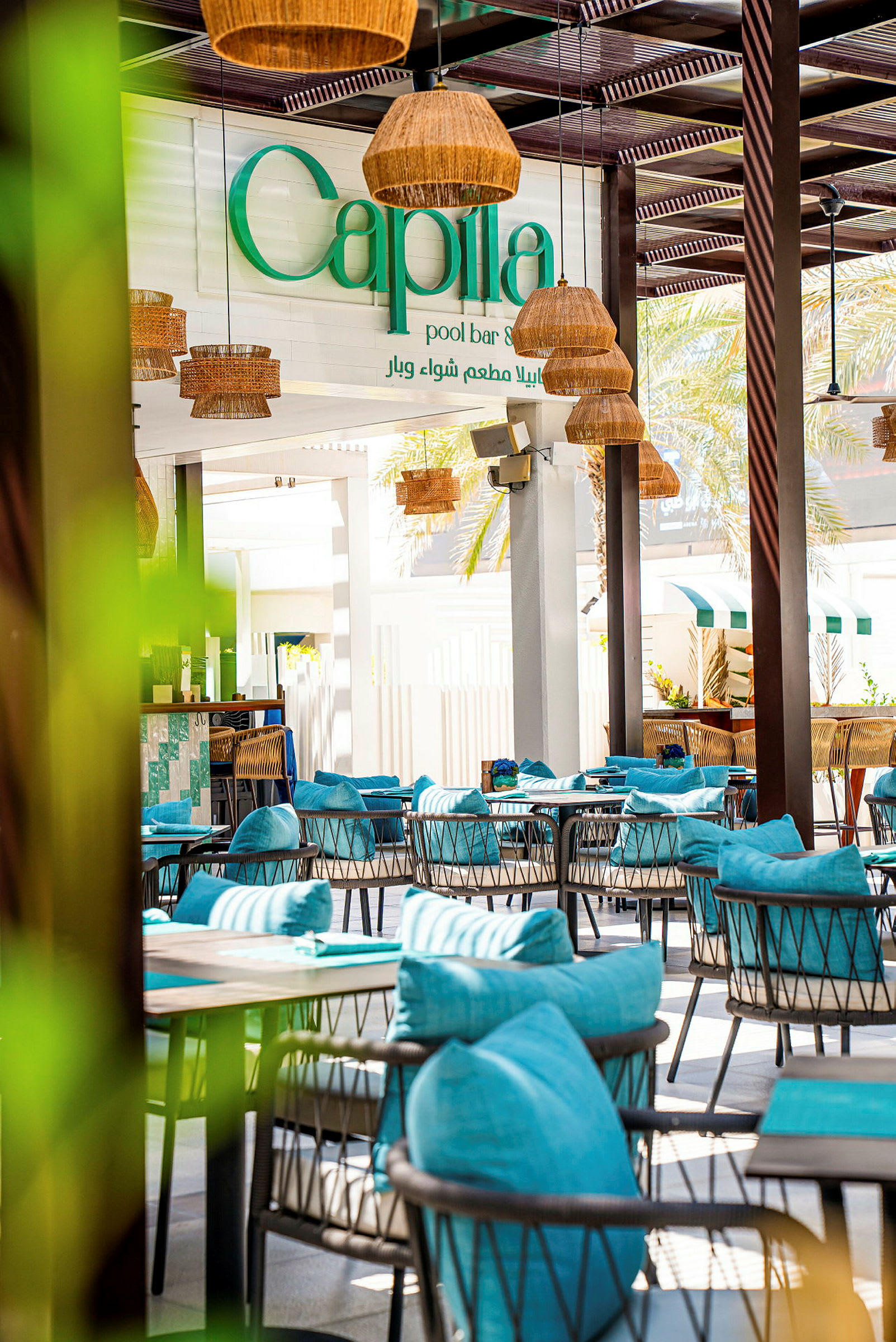 En utomhus bar och restaurang med turkosa sittmöbler och dekorerad med rottinglampor. En skylt med texten "Capila pool bar" syns i bakgrunden. (AI-genererad text)