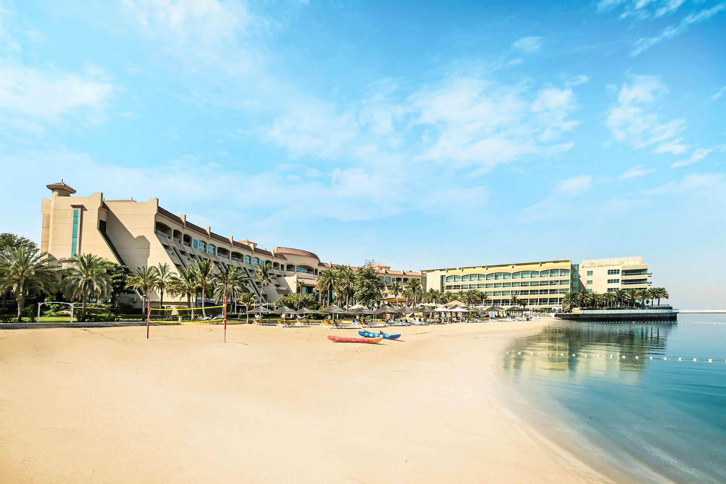 Hotellbyggnad vid en sandstrand med palmer, solstolar och spegelblankt vatten. (AI-genererad text)