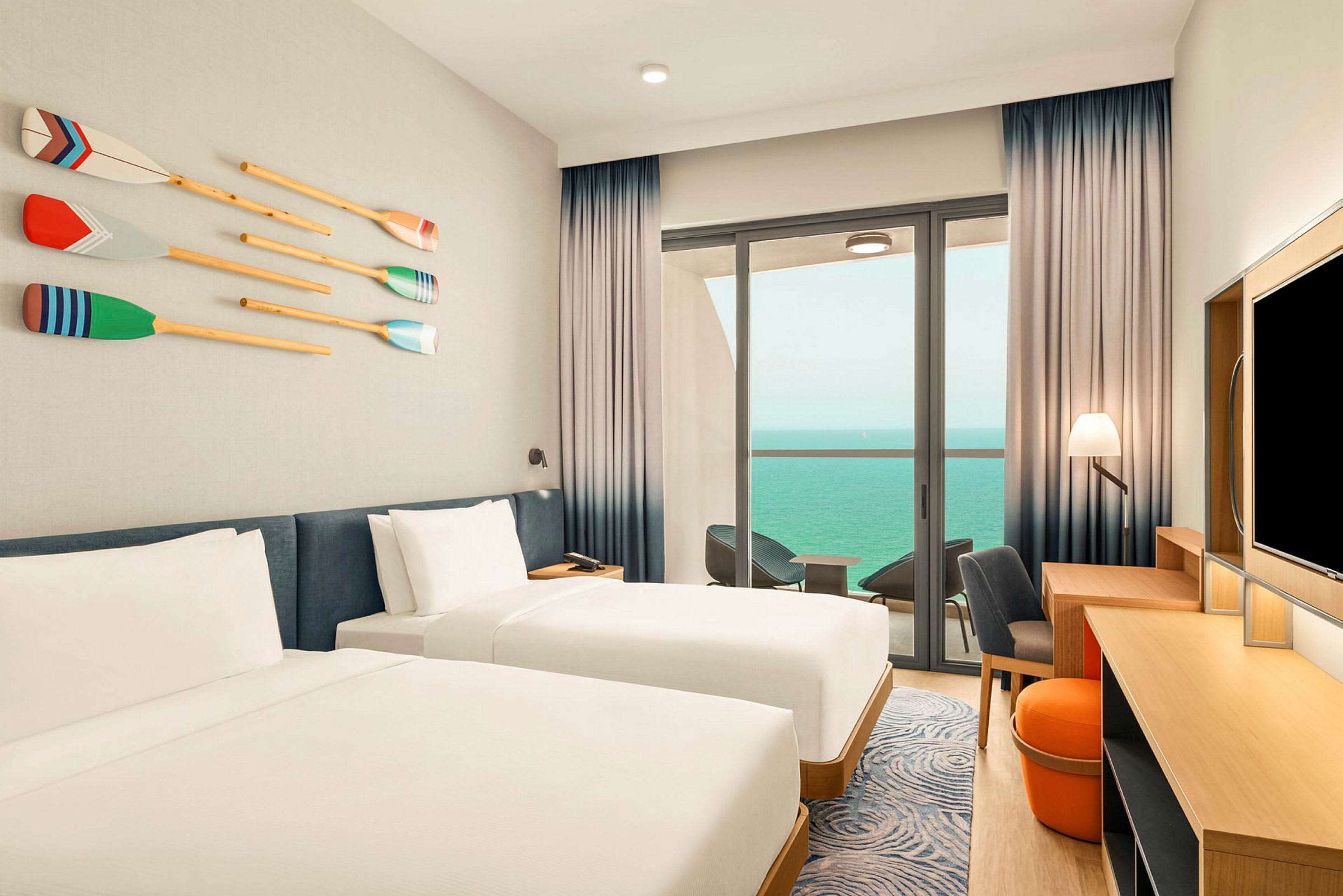 Ett hotellrum med två enkelsängar, en väggdekoration föreställande pinnar, och en balkong med utsikt över havet. (AI-genererad text)