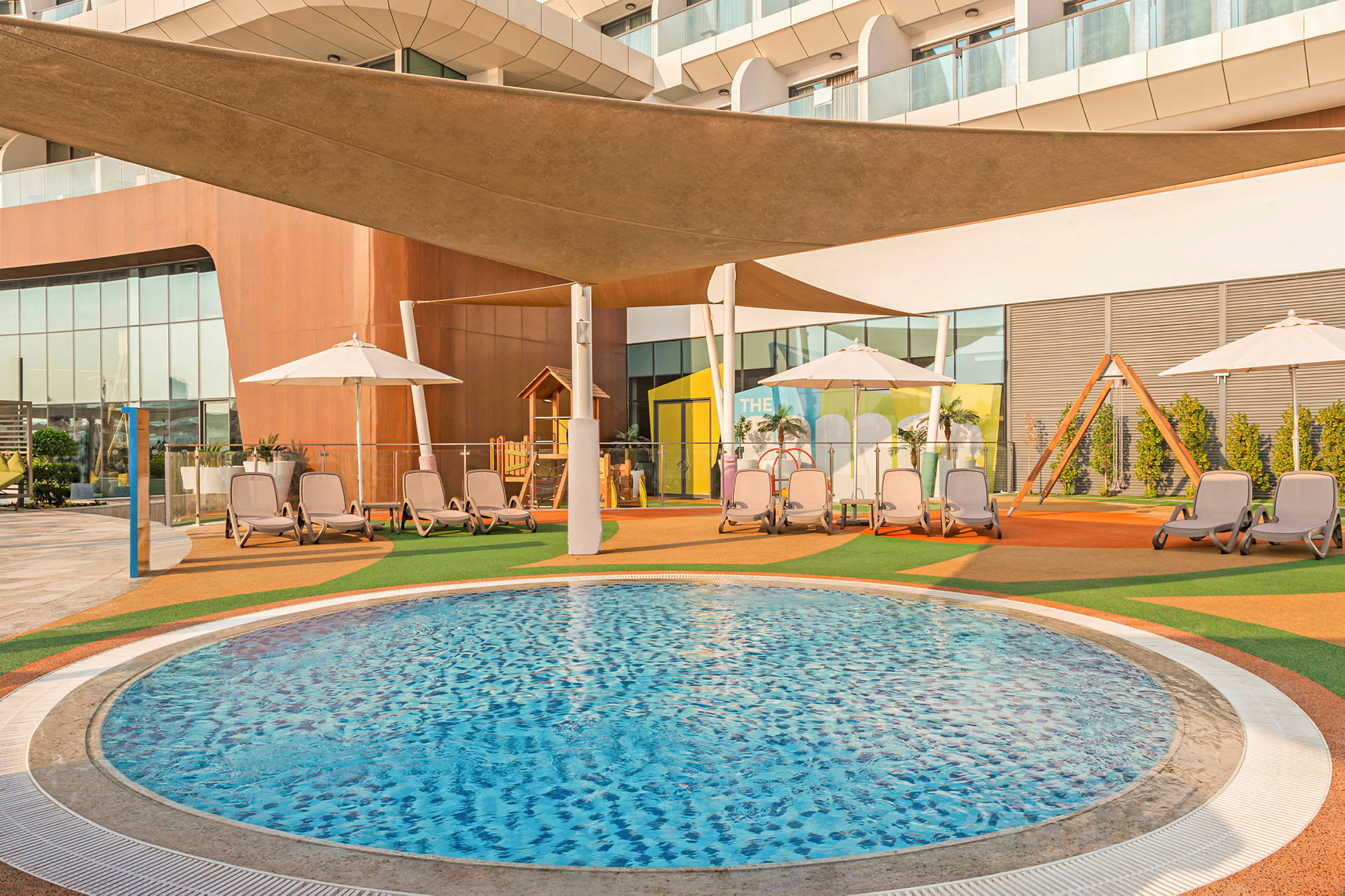 Bild av en rund pool i ett utomhusområde med solstolar, parasoller, en lekplats och moderna byggnader i bakgrunden. (AI-genererad text)