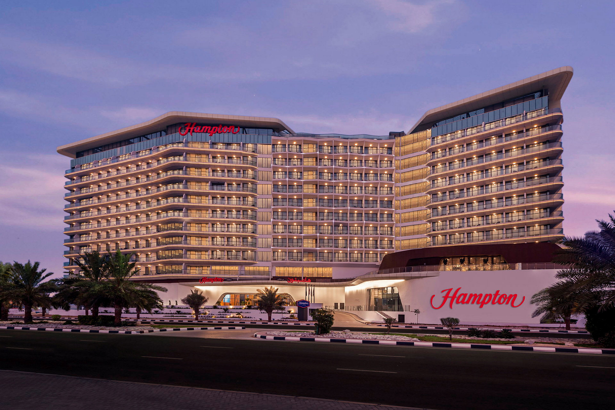Ett stort hotell med namnet "Hampton" syns i kvällsljuset. Byggnaden har flera våningar med många balkonger och palmer pryder framsidan. (AI-genererad text)
