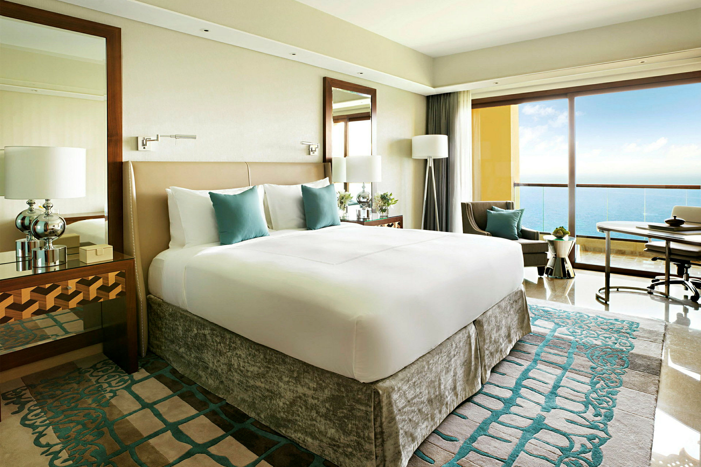 Ett modernt hotellrum med en stor säng, vita och turkosa sängkläder, och en balkong med utsikt över havet. (AI-genererad text)