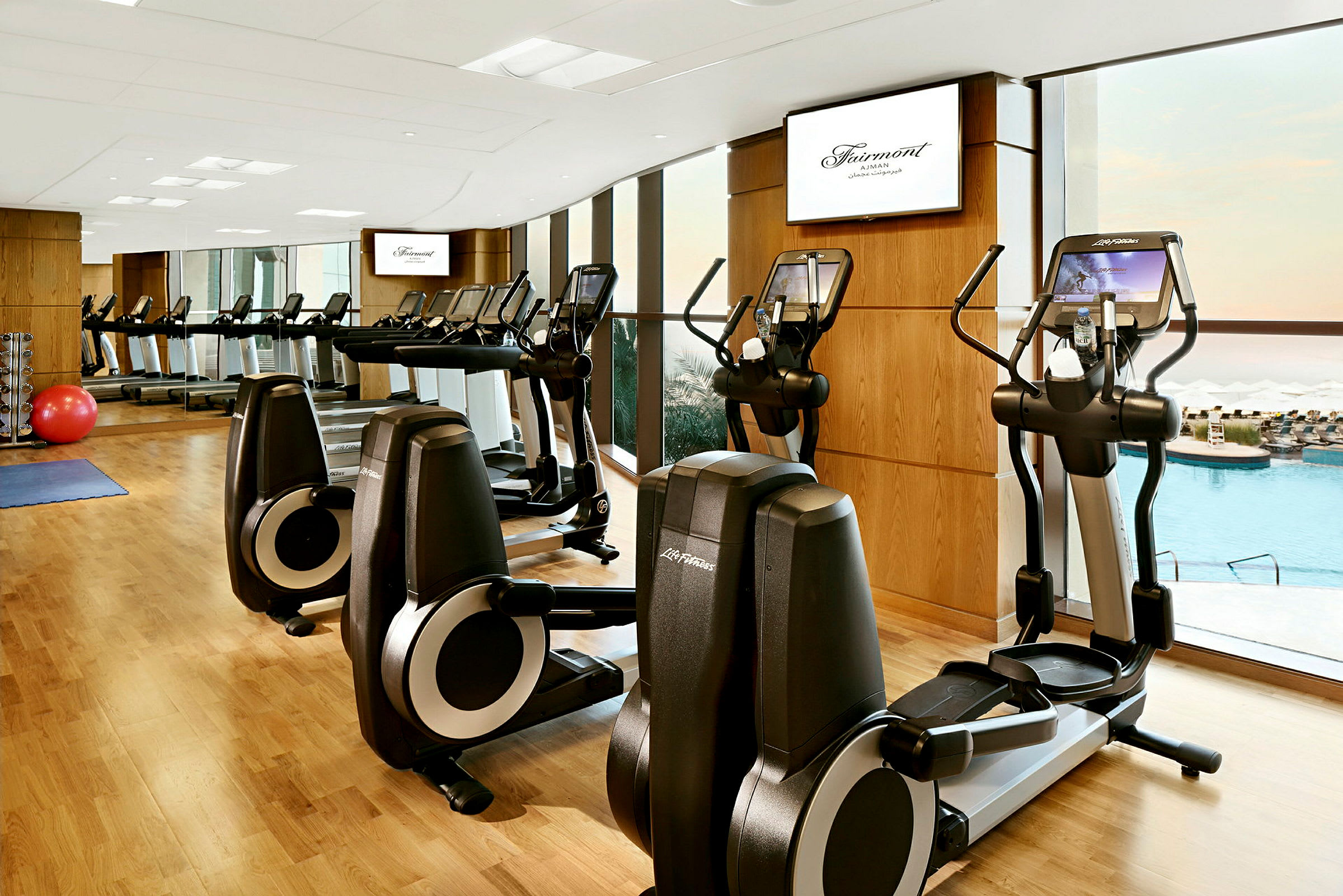 Ett gym med träningsmaskiner, träväggar, speglar och utsikt mot en pool genom stora fönster. (AI-genererad text)