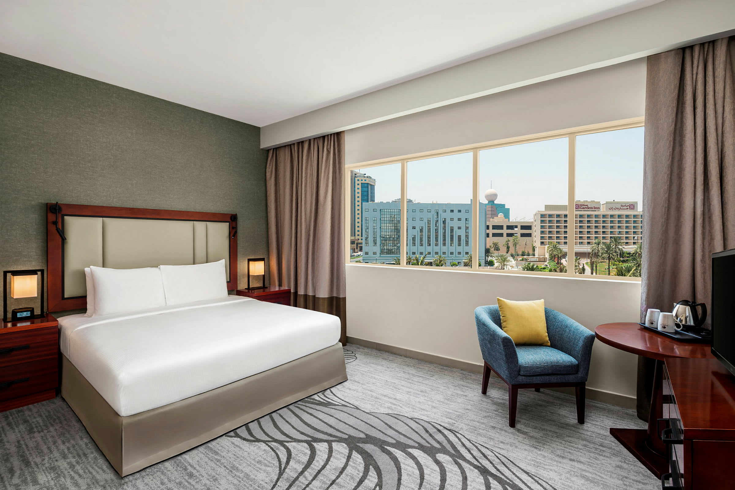 Ett hotellrum med en stor säng, sittmöbler, ett skrivbord och ett fönster som visar en stadsvy utanför. (AI-genererad text)