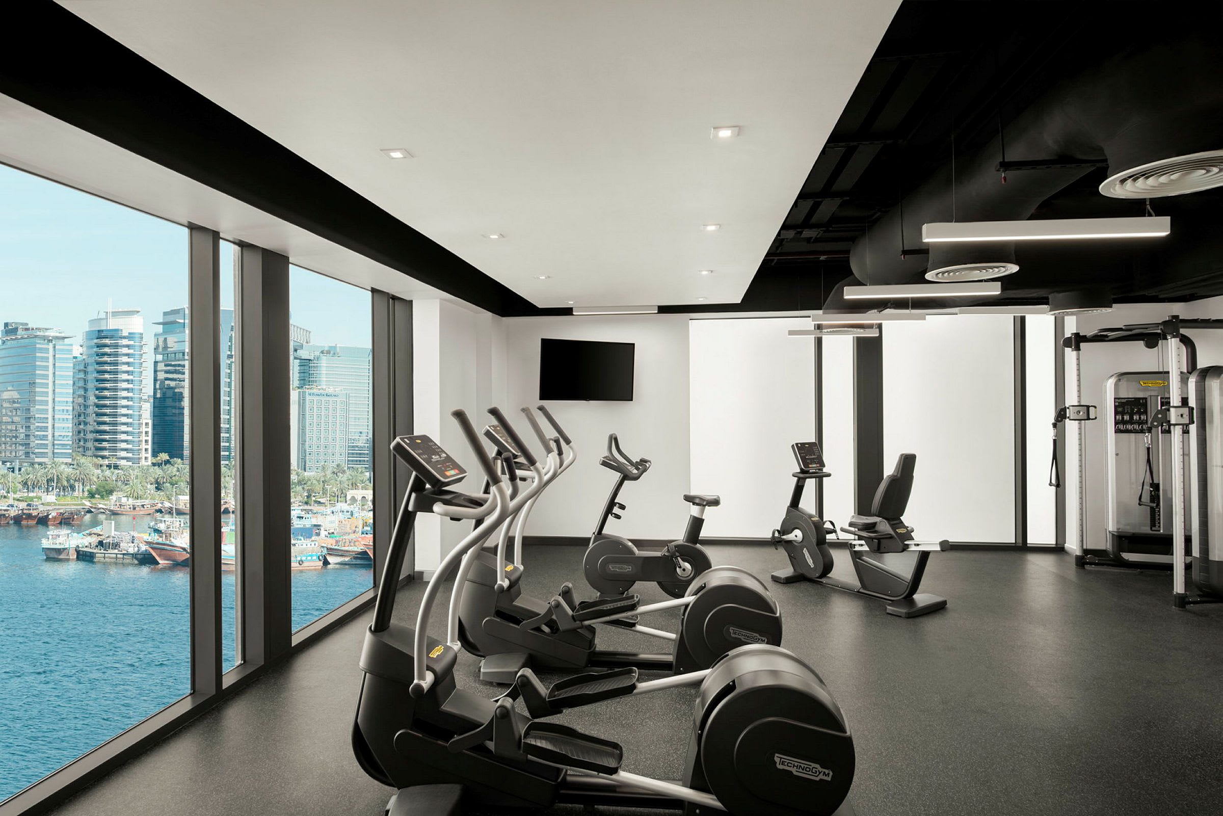 En modern gymmiljö med träningsmaskiner och stora fönster som vetter mot en utsikt av vattnet och stadssilhuetten. (AI-genererad text)