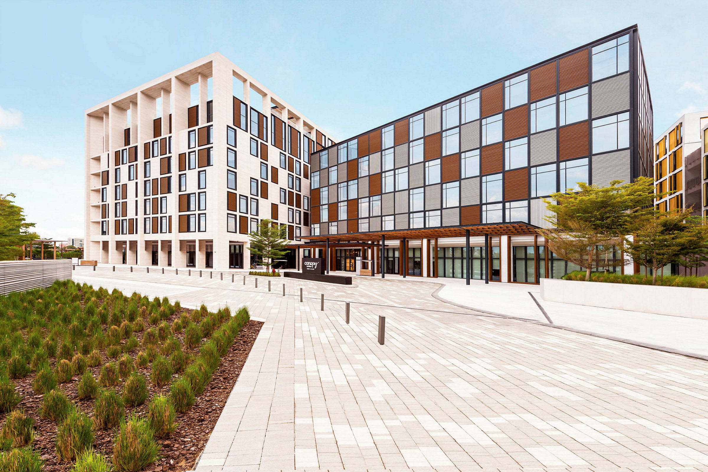 Ett modernt hotell med en ljus fasad och stora fönster, beläget vid ett öppet torg. (AI-genererad text)
