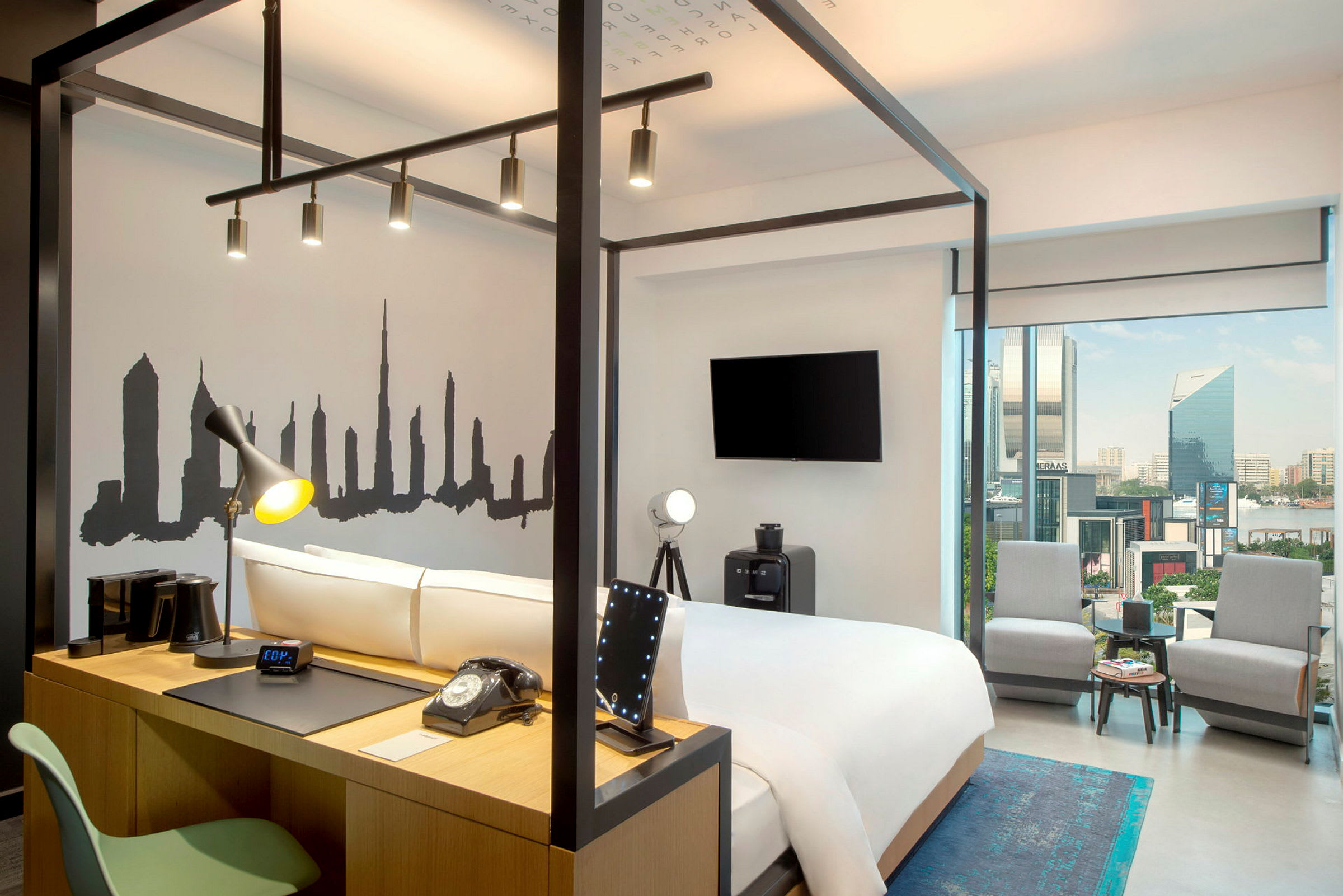 Ett modernt hotellrum med en säng omgiven av en stålram, en väggdekoration föreställande en stadssilhuett, och ett skrivbord med lampa och telefon. Det finns stora glasfönster som erbjuder utsikt över staden. (AI-genererad text)