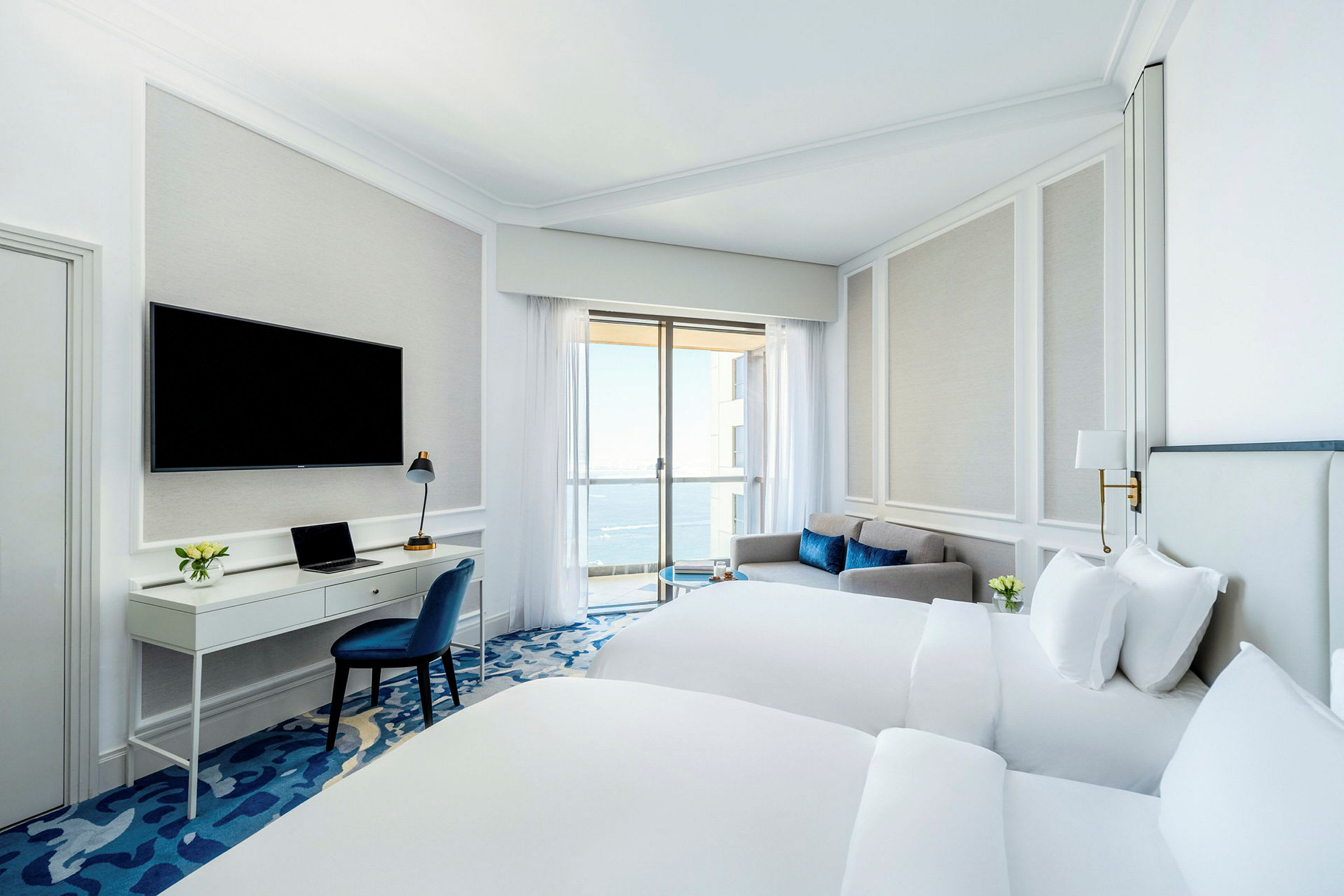 Ett hotellrum med två sängar, en arbetsyta med en stol, en soffa och en balkong med havsutsikt. (AI-genererad text)