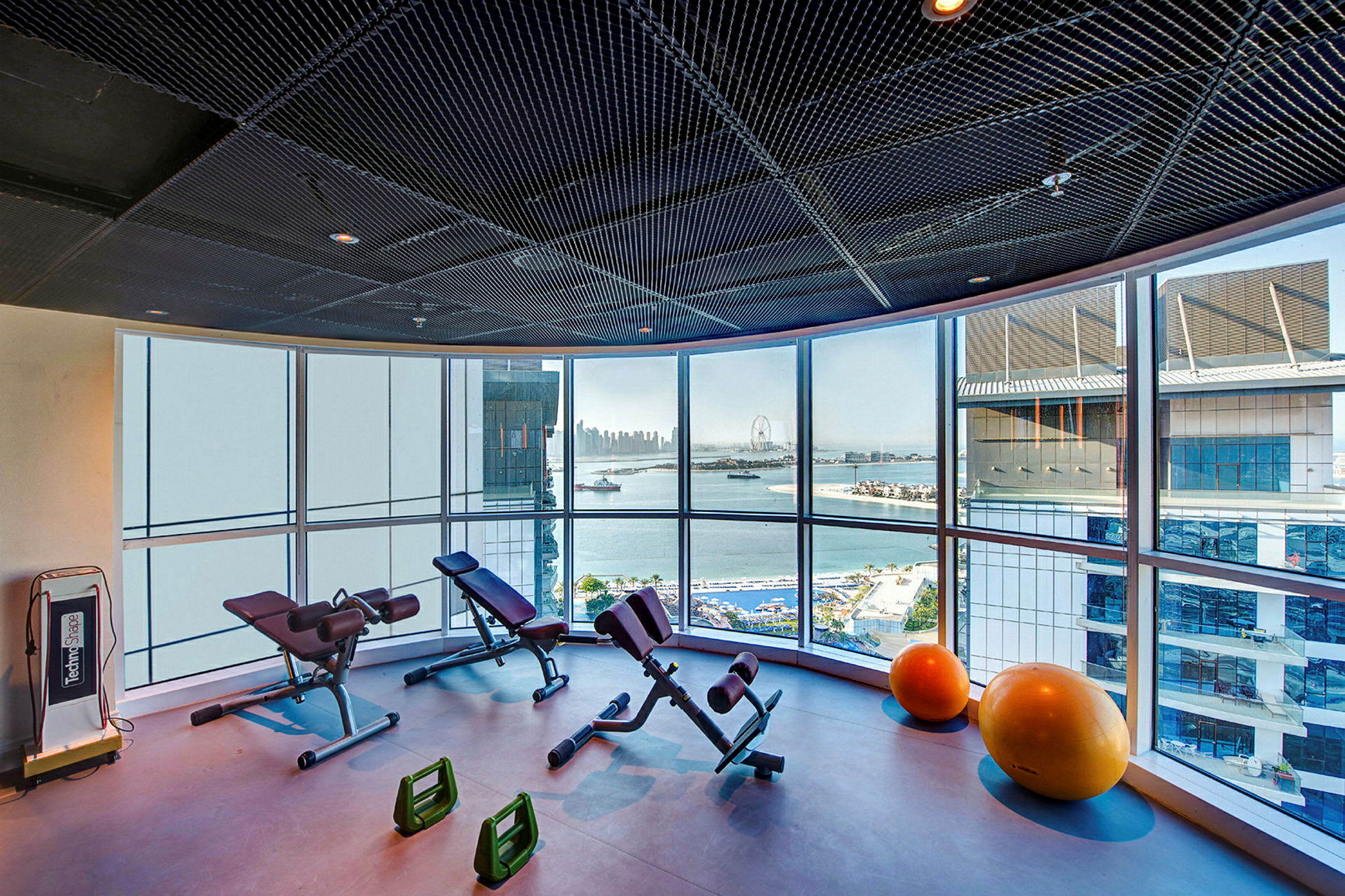 Ett gym med träningsutrustning och stora fönster som visar utsikt över havet och stadsmiljö. (AI-genererad text)