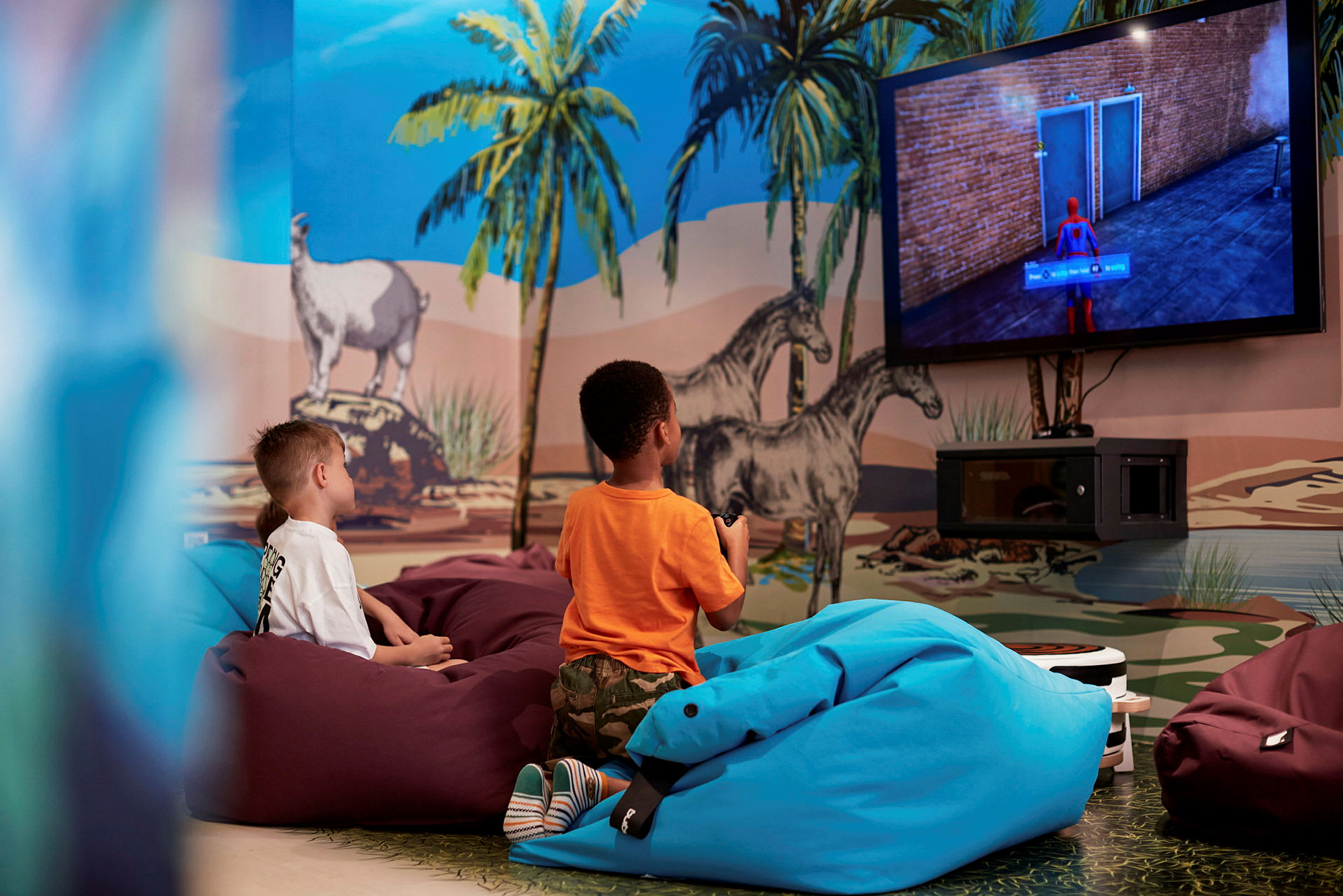 Två barn sitter i en färgglad lekhörna med en väggmålning av djur och natur, medan de tittar på en TV. (AI-genererad text)