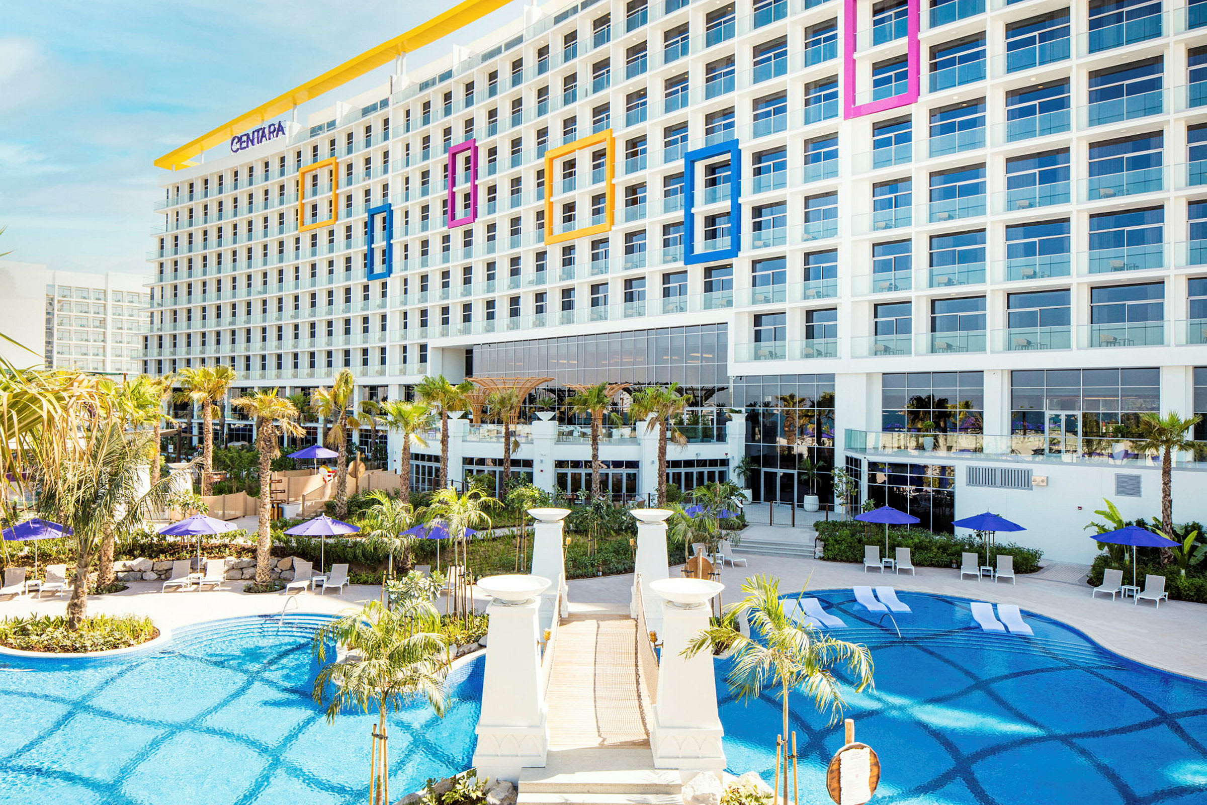 Ett hotell i modern stil med stora fönster, omgiven av palmer och en pool med solstolar och parasoller. (AI-genererad text)