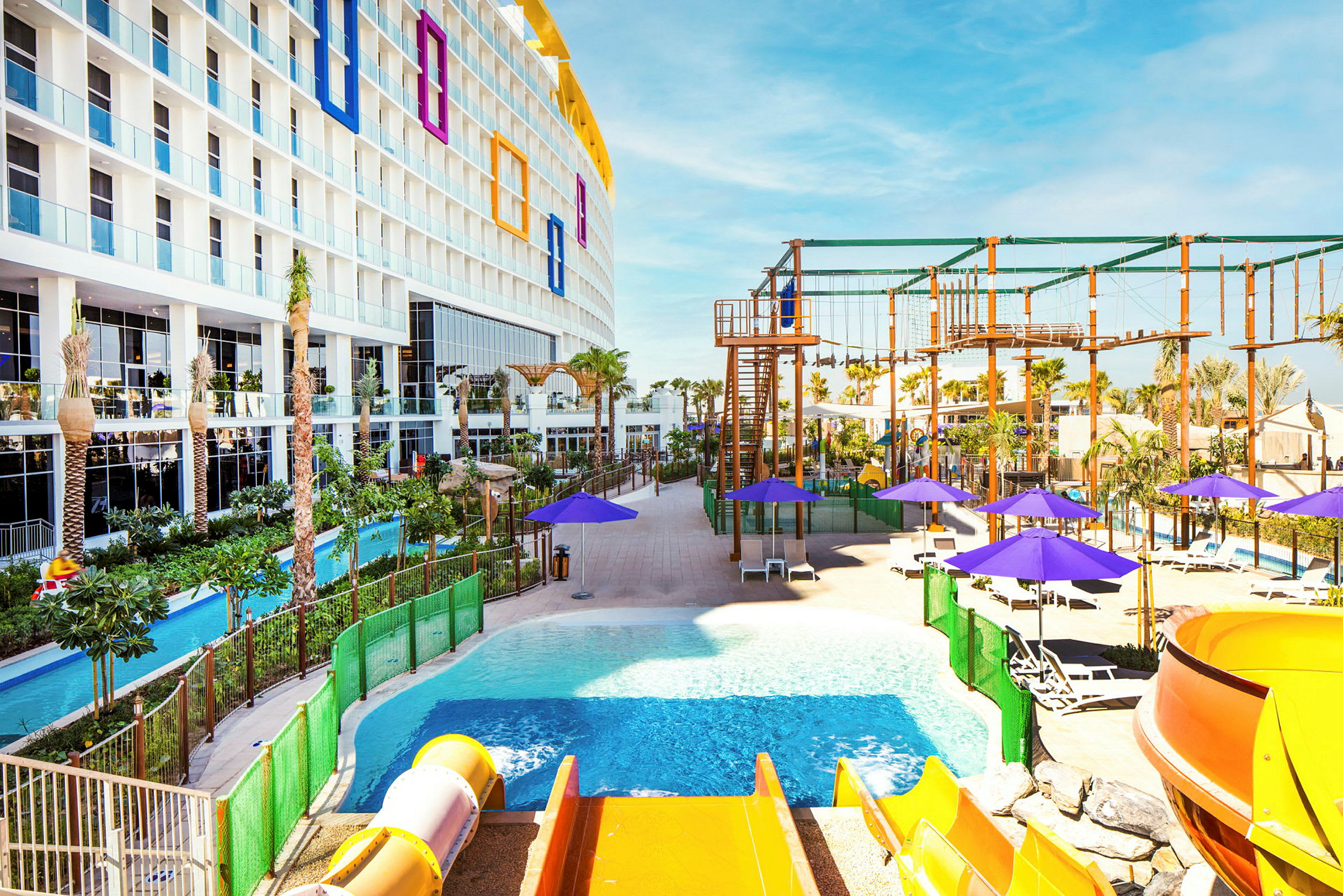 Ett hotell med flera våningar och balkonger med utsikt över ett poolområde med en vattenrutschkana, solstolar och parasoller. I bakgrunden syns ett klätterställningsområde och grönska. (AI-genererad text)