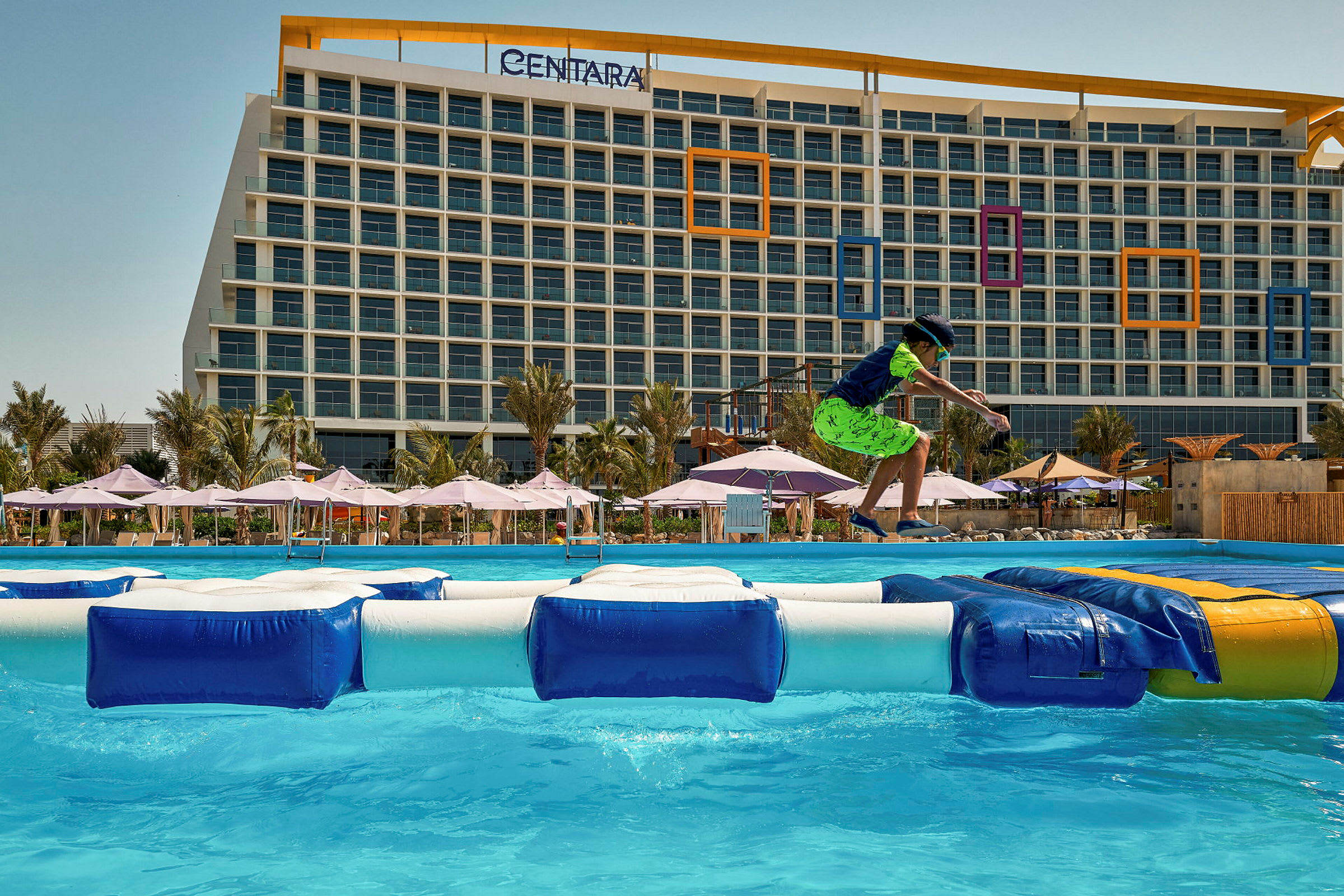 En stor hotellbyggnad med parasoller vid en pool. En person leker på uppblåsbara leksaker i vattnet. (AI-genererad text)