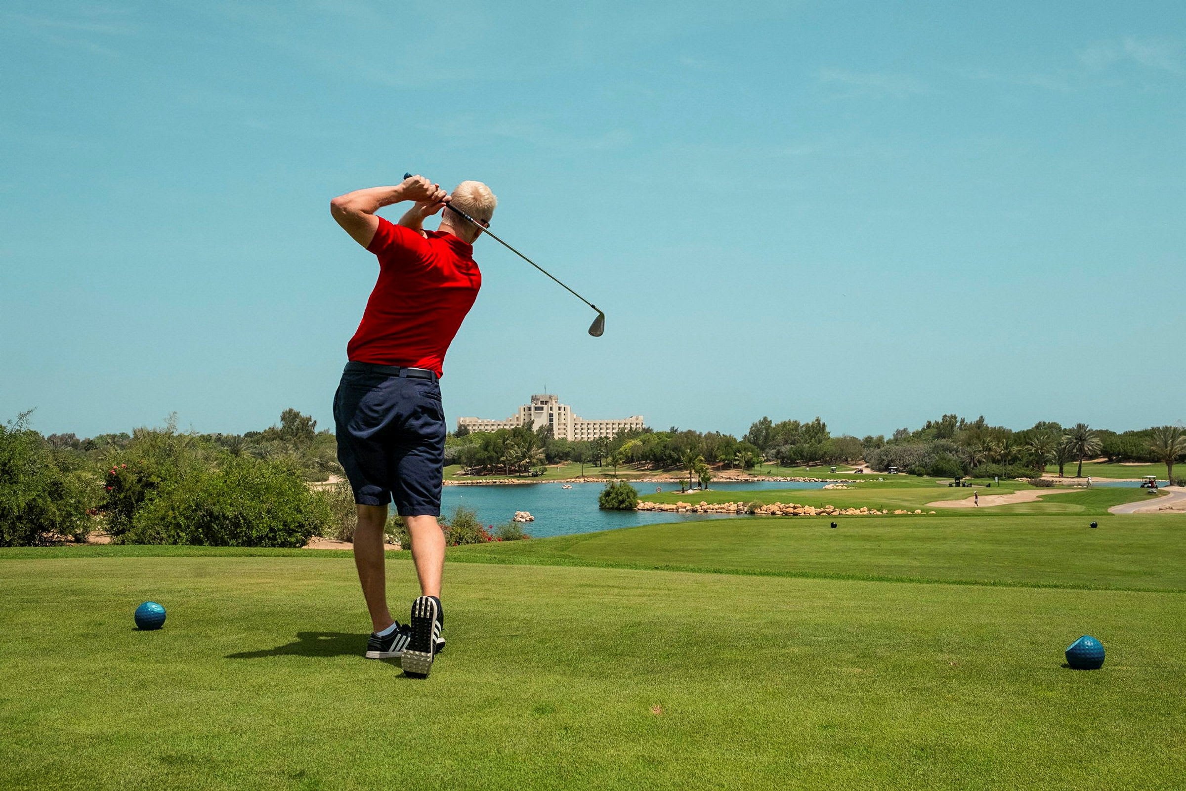 En person klädd i rött spelar golf på en bana med grön gräsmatta, omgiven av träd och vatten. Ett hotell syns i bakgrunden. (AI-genererad text)