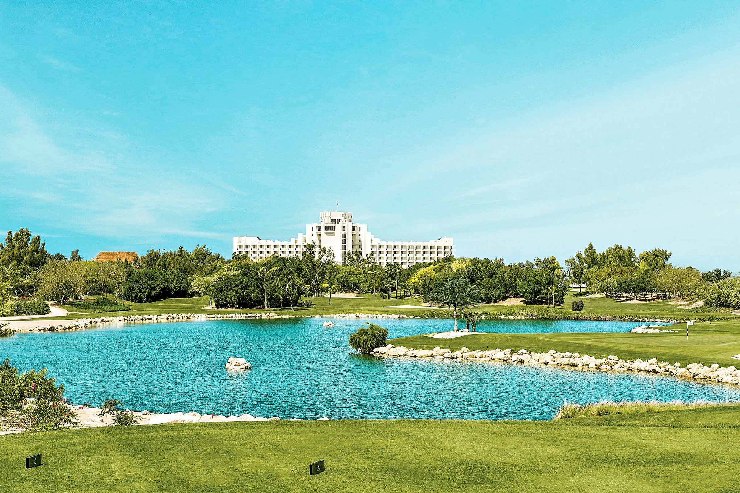 Vy över en golfbana med en damm i förgrunden och ett stort vitt hotell i bakgrunden. (AI-genererad text)