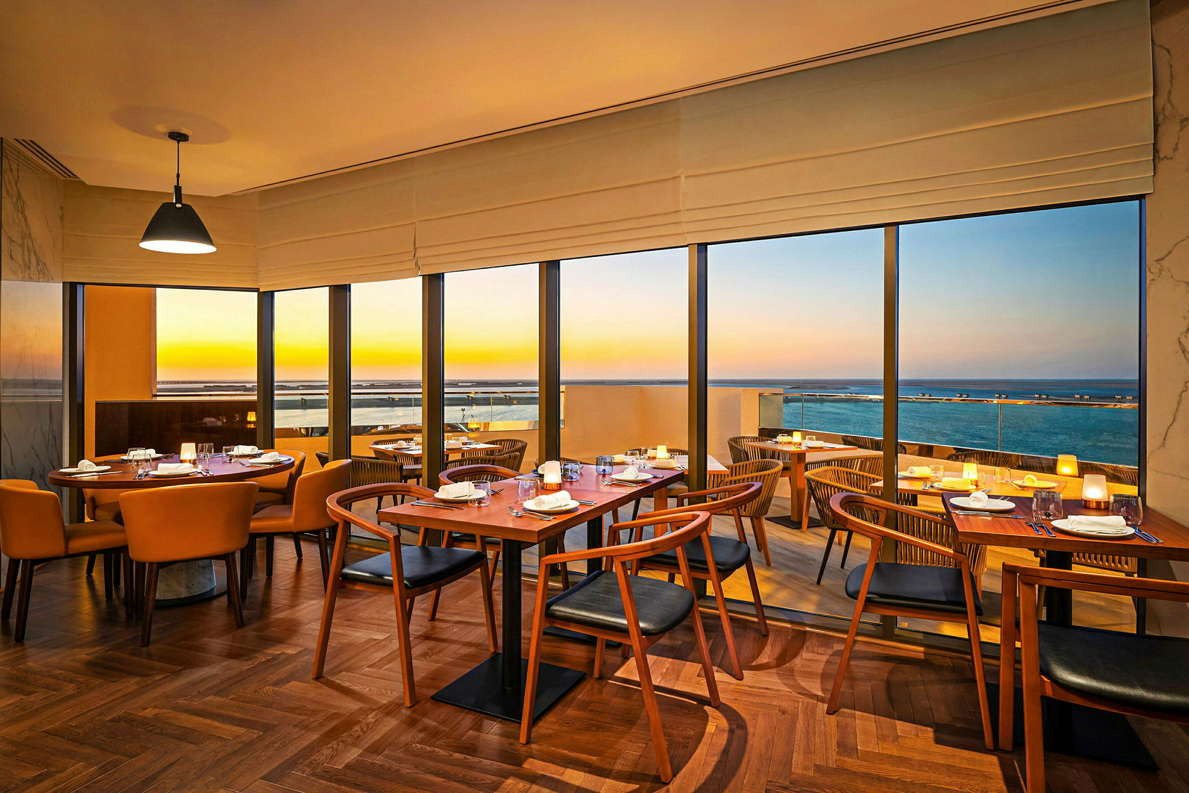 Interiör av en restaurang med bord och stolar samt utsikt över havet i solnedgång. (AI-genererad text)