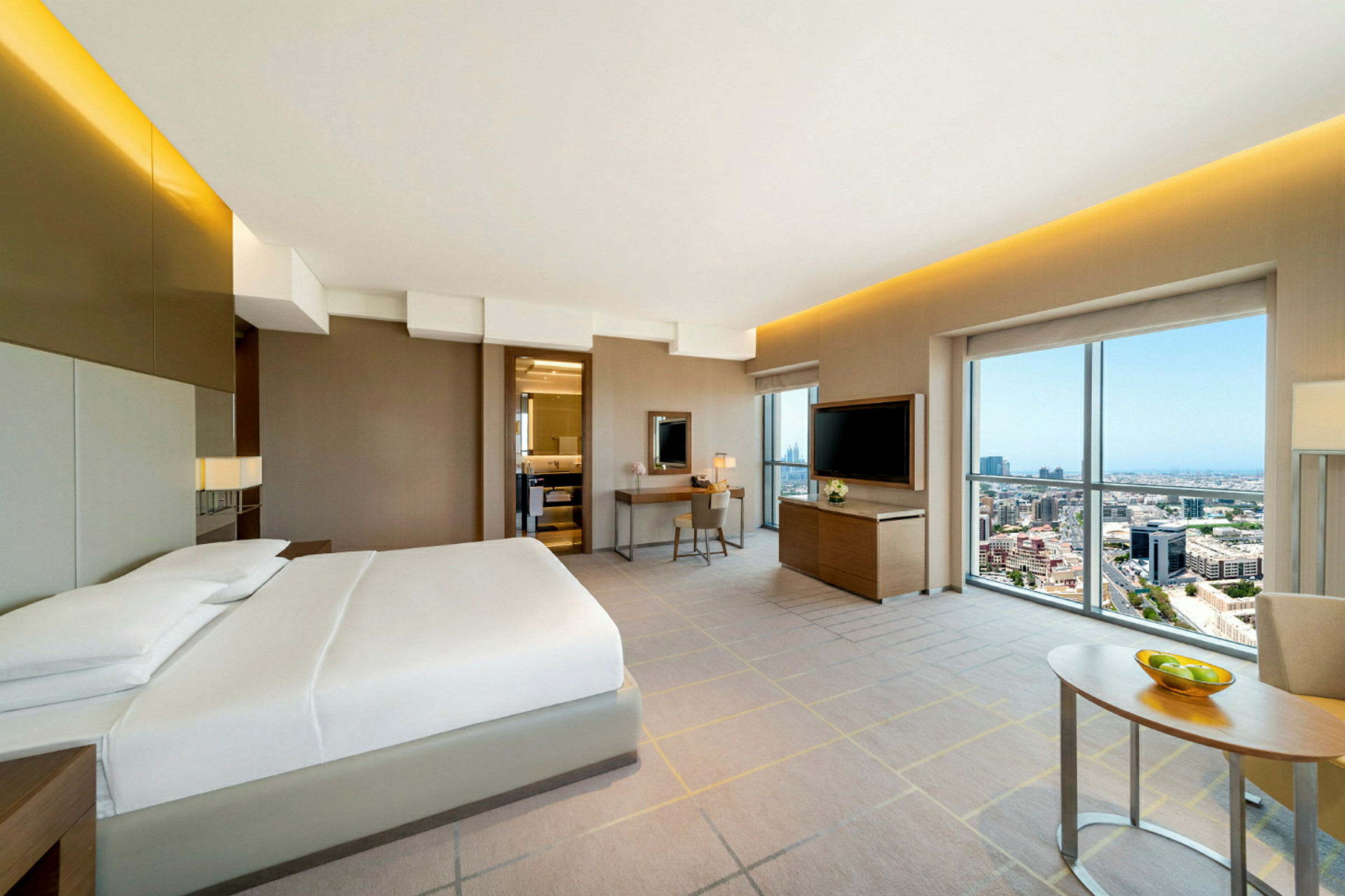 Interiör av ett hotellrum med dubbelsäng, skrivbord, TV och stort fönster med utsikt över en stad. (AI-genererad text)