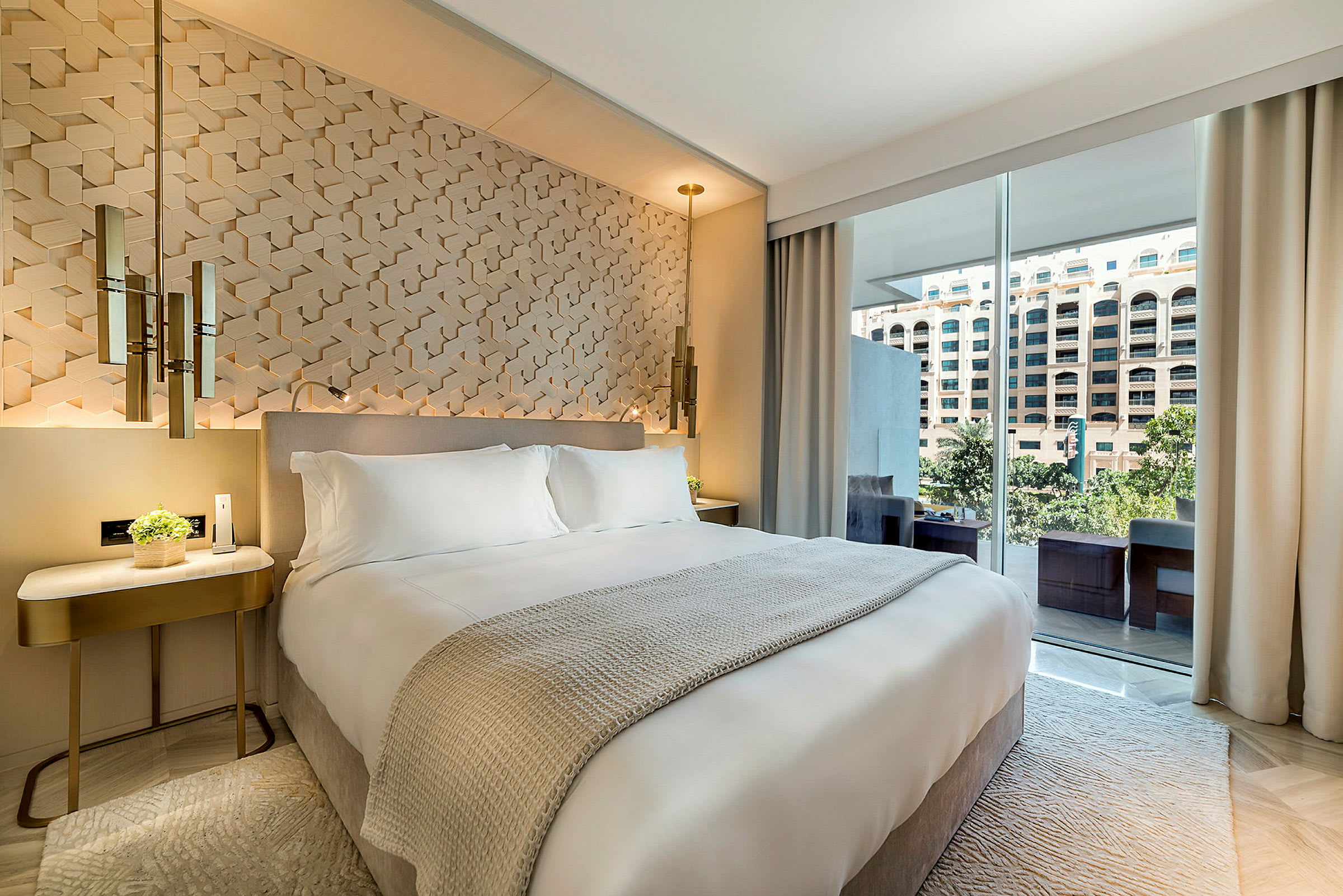 Bild på ett elegant hotellrum med en stor säng, designad väggdekoration bakom sängen och stora fönster som leder ut till en balkong med utsikt mot en byggnad. (AI-genererad text)